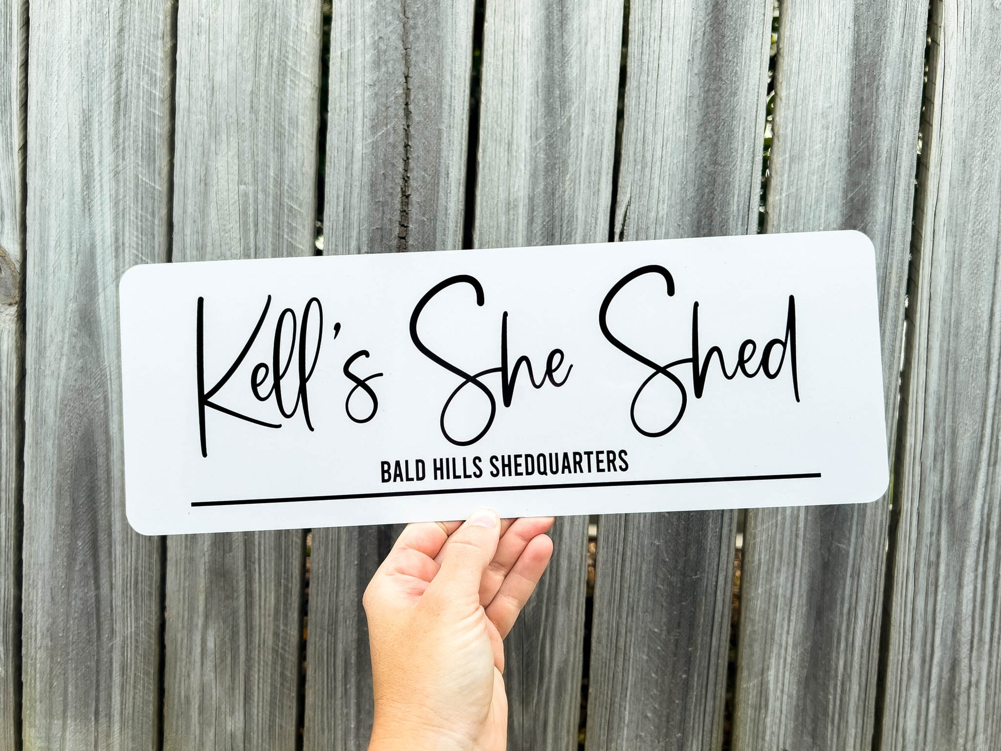 Hand holding a white sign with 'Kell's She Shed' in black text against a wooden background