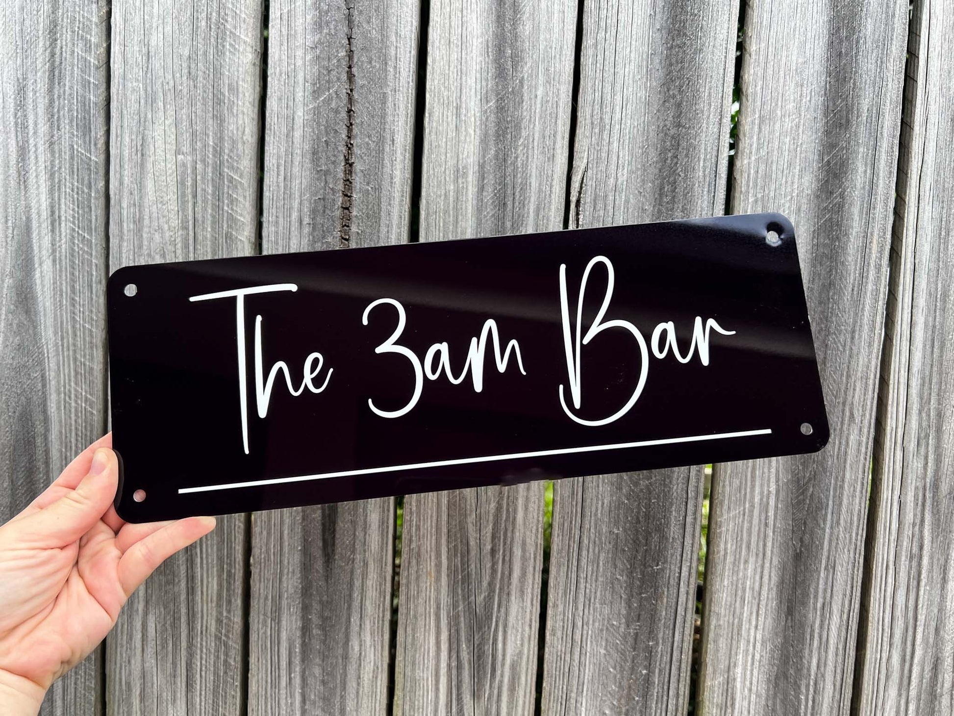 Hand holding a personalised black and white sign with 'The 3am Bar' text against a wooden fence background