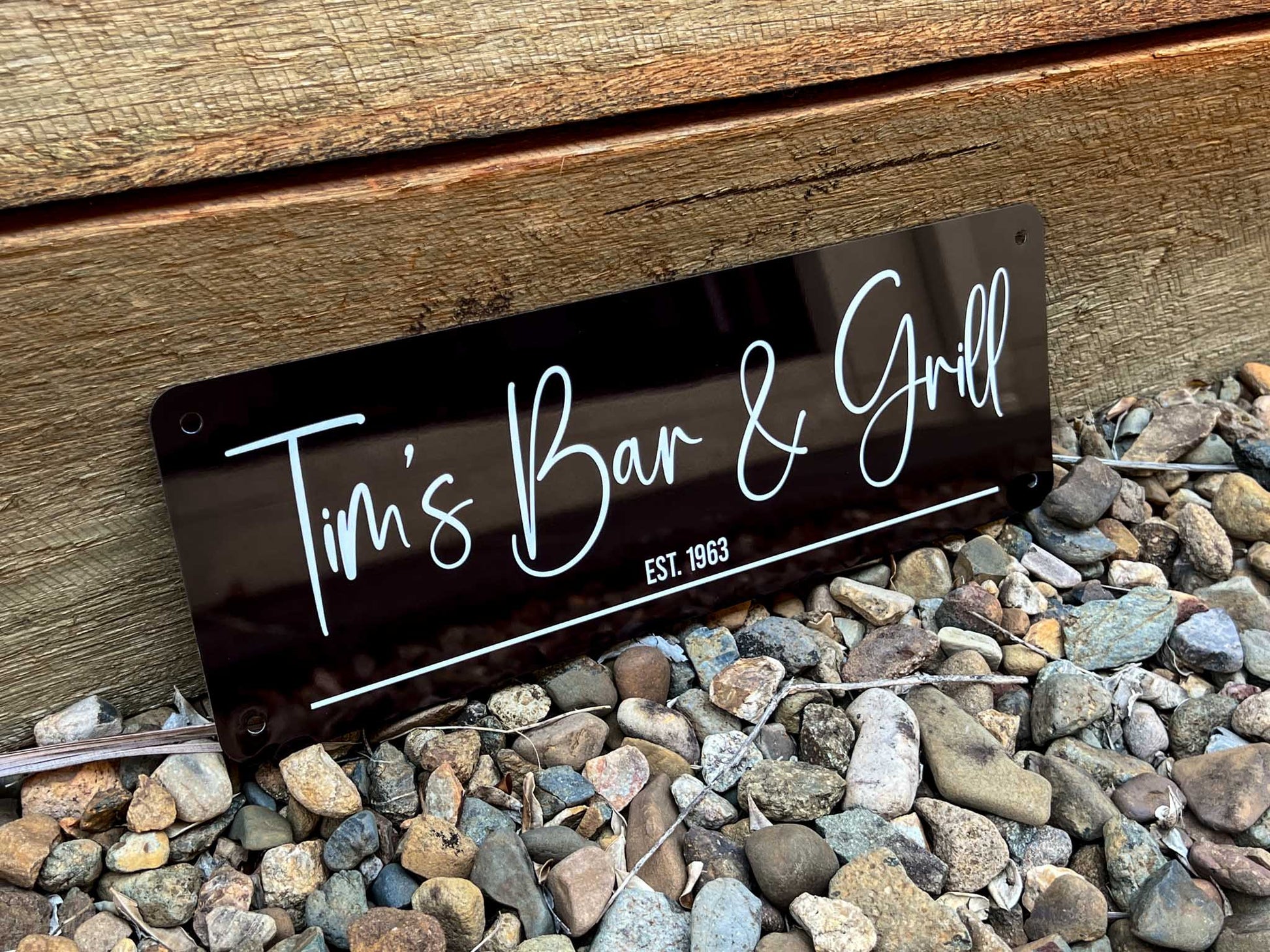 A custom printed metal sign reading 'Tim's Bar & Grill EST. 1983' on a wooden surface with pebbles below.