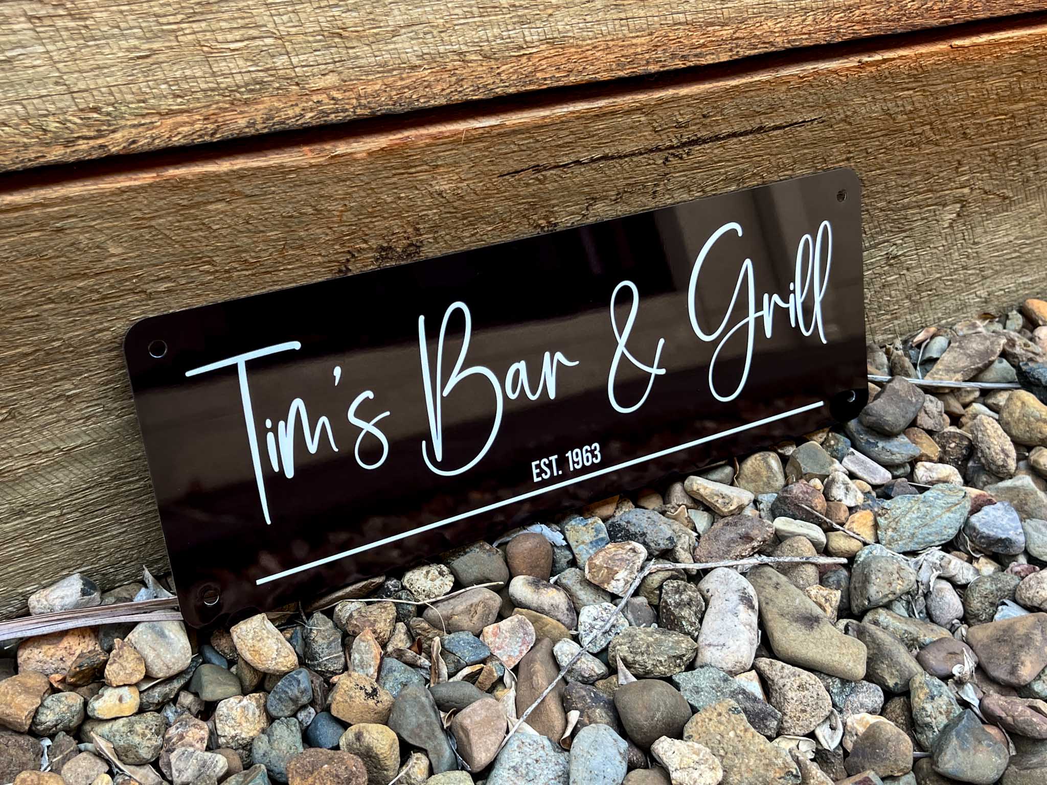 A custom printed metal sign reading 'Tim's Bar & Grill EST. 1983' on a wooden surface with pebbles below.