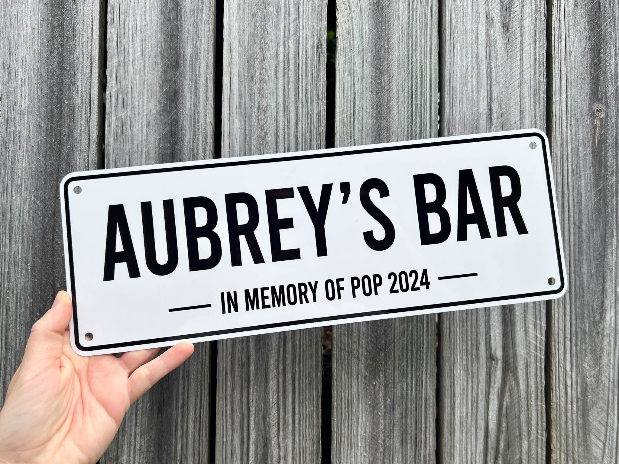 Hand holding a bar sign that reads 'Aubrey's Bar In Memory of Pop 2024' against a wooden background.