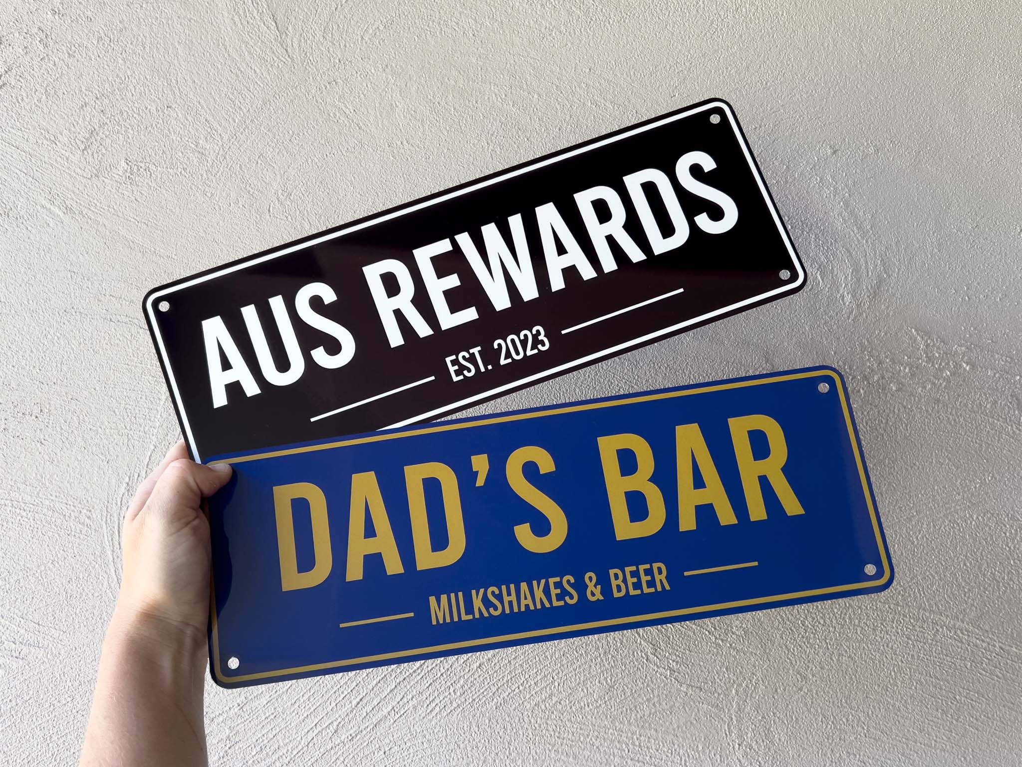 Two custom printed bar signs, one black with 'AUS REWARDS' and another blue with 'DAD'S BAR', held against a light background.
