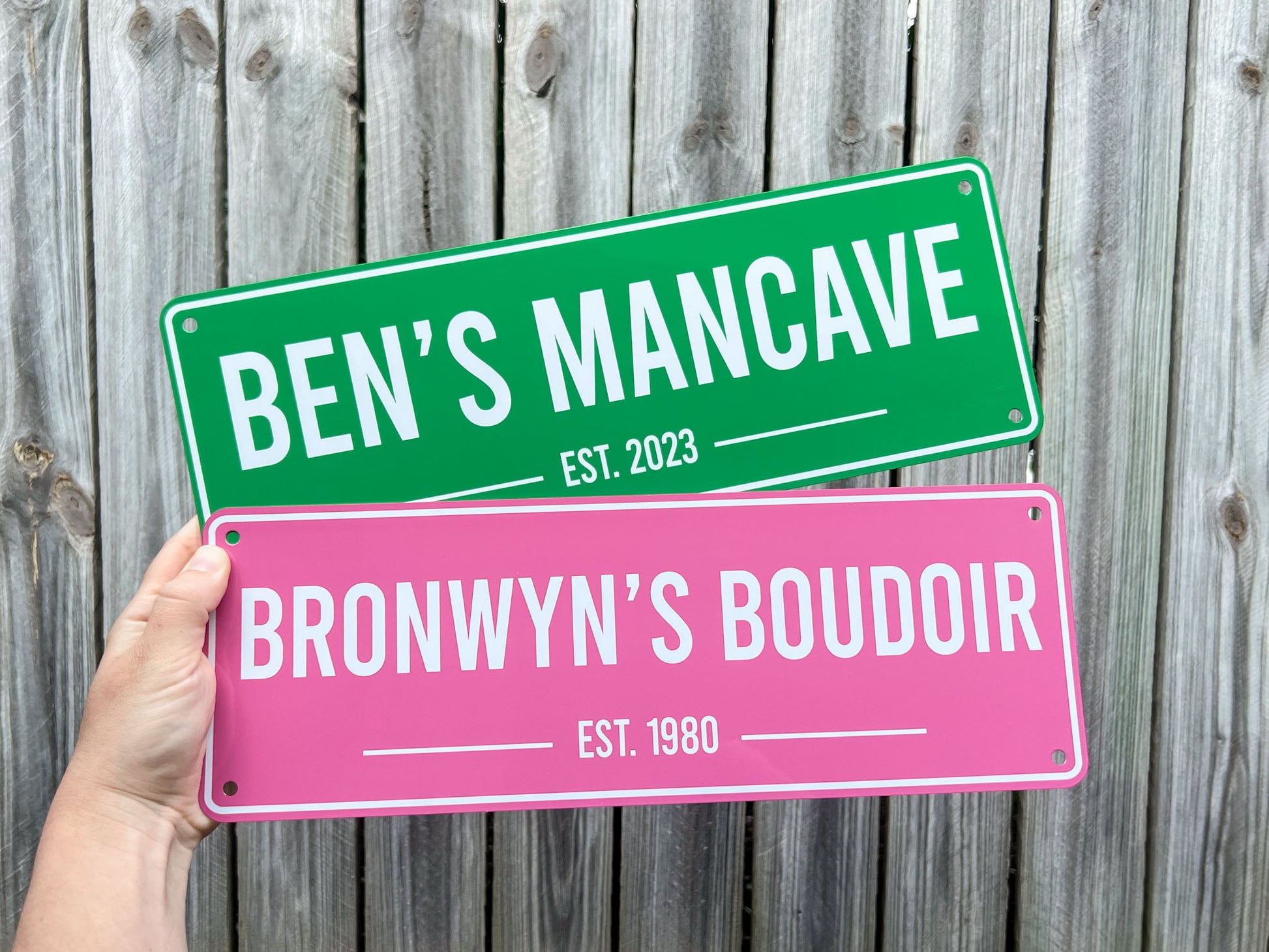 Two personalized bar signs in apple green and pink held against a wooden fence background.