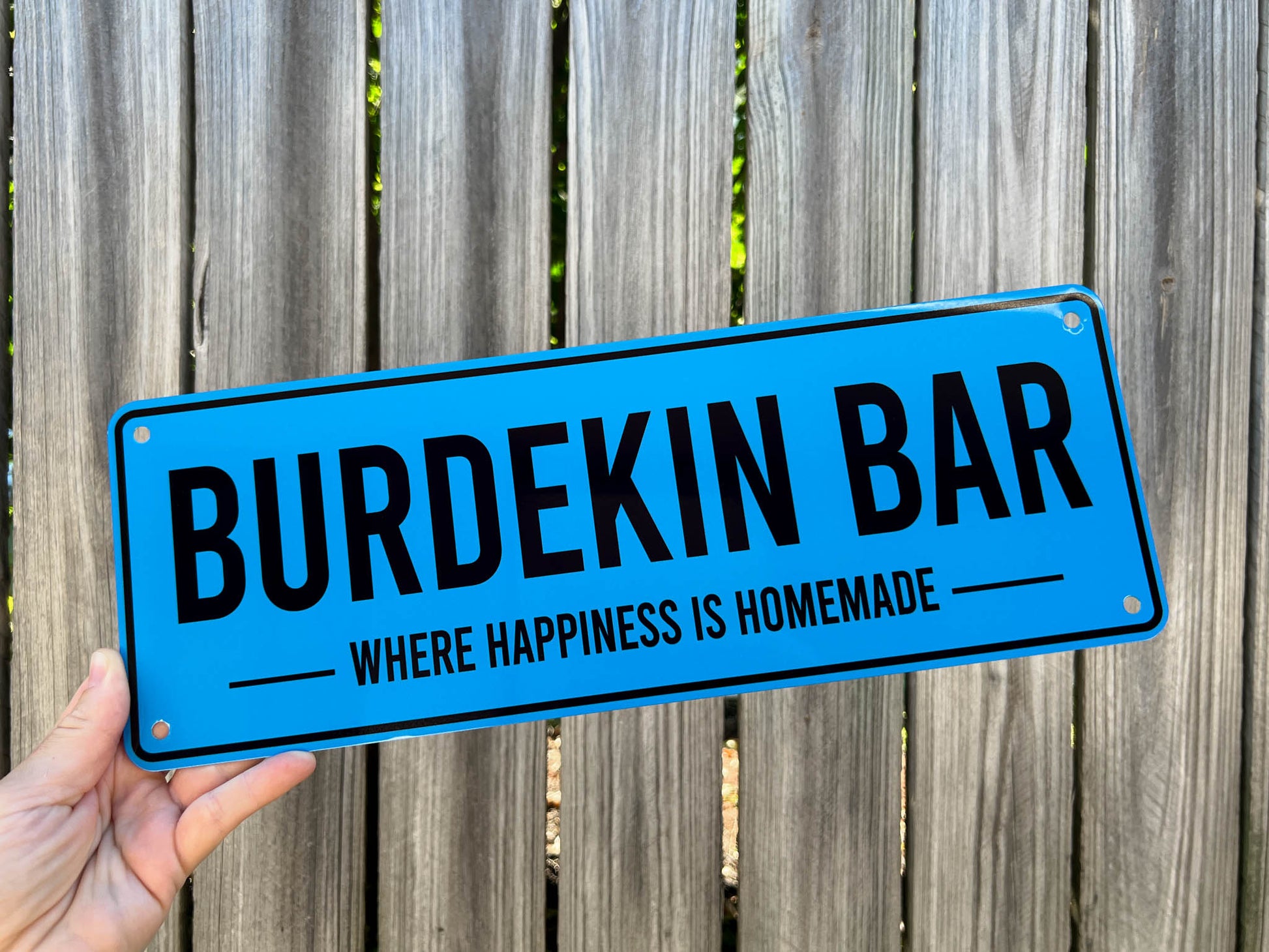 Blue sign with 'Burdekin Bar' and 'Where Happiness is Homemade' text held against a wooden fence.