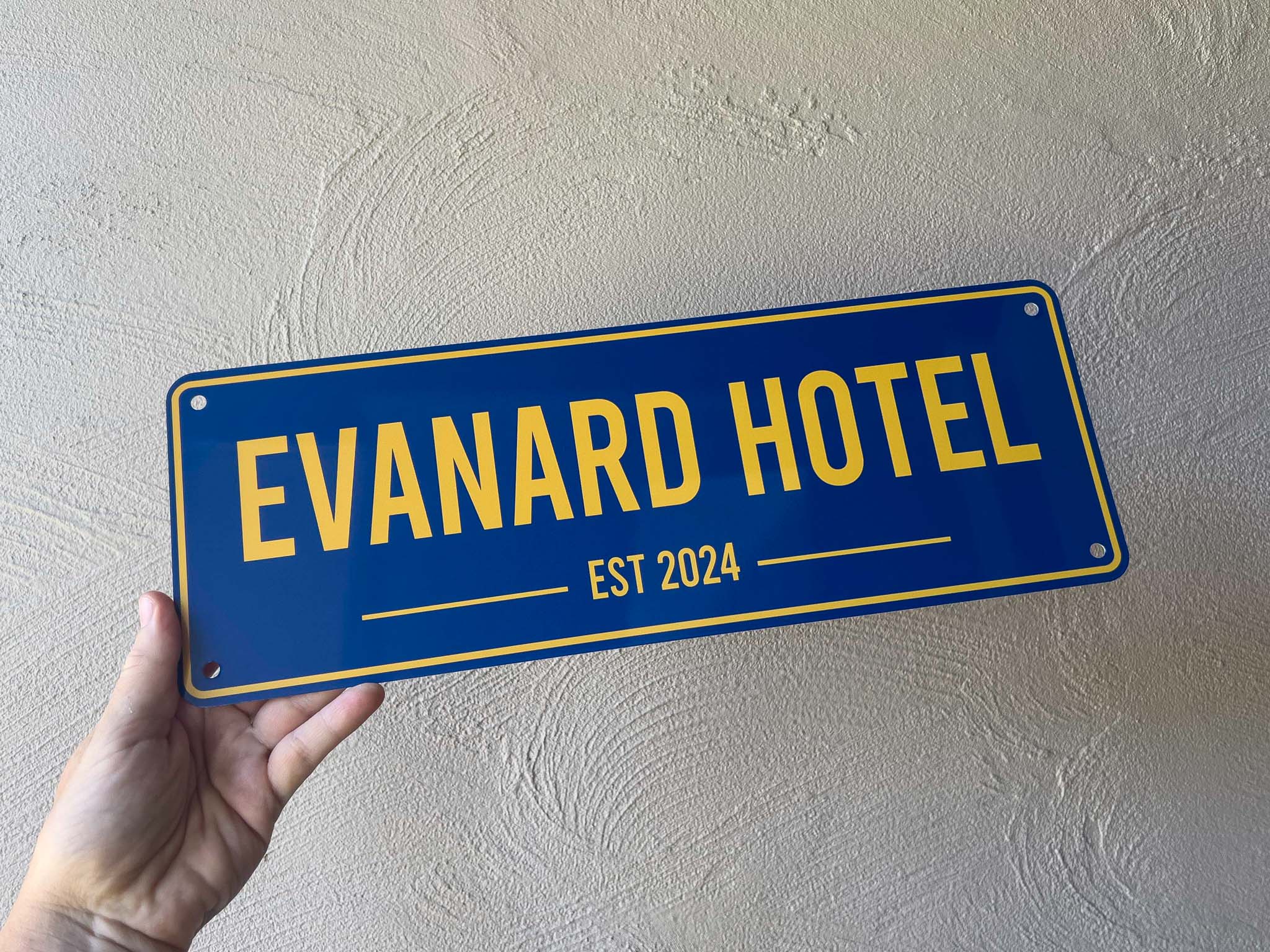 Hand holding a blue sign with 'Evanard Hotel' and 'Est 2024' text against a textured wall.