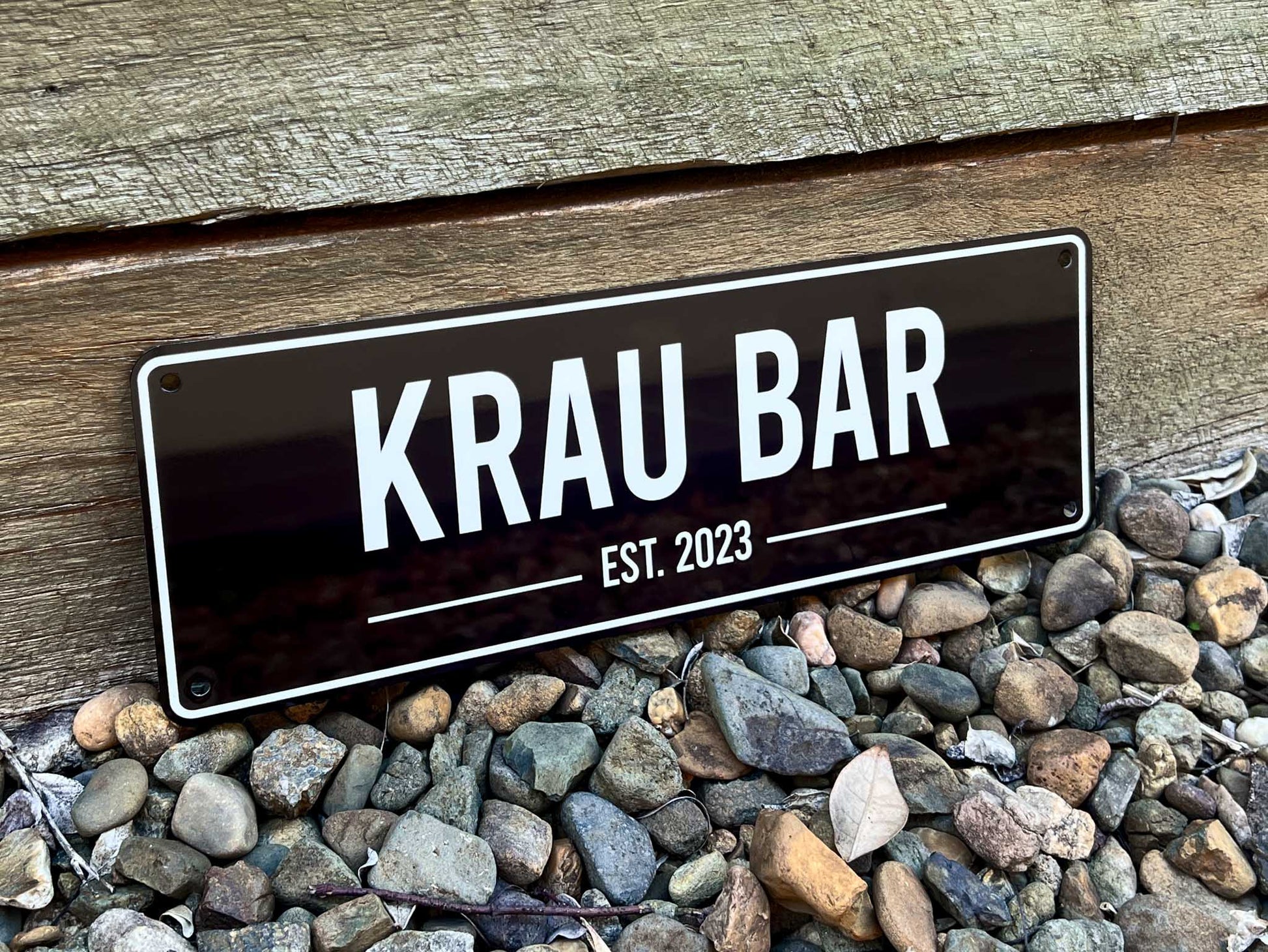 A personalised metal sign with 'KRAU BAR EST. 2023' on a wooden surface and pebbles.