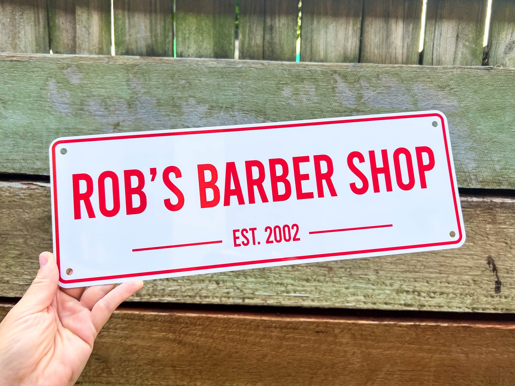 Hand holding a sign that reads 'Rob's Barber Shop EST. 2002' against a wooden background.