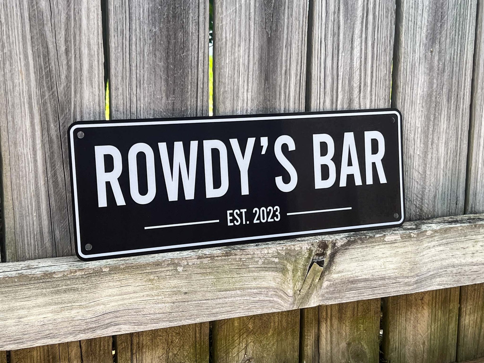 Black sign with 'ROWDY'S BAR EST. 2023' text on a wooden background