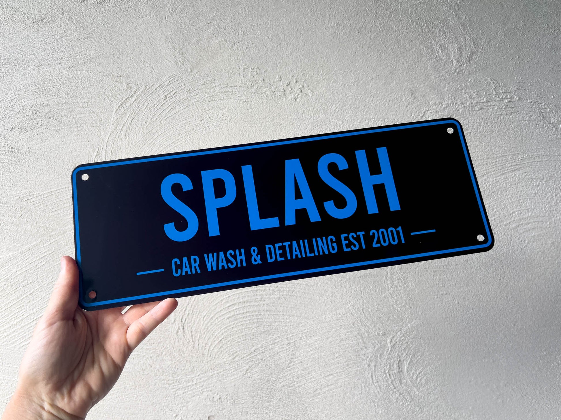Hand holding a blue and black sign with 'SPLASH Car Wash & Detailing Est 2001' text against a light gray background.