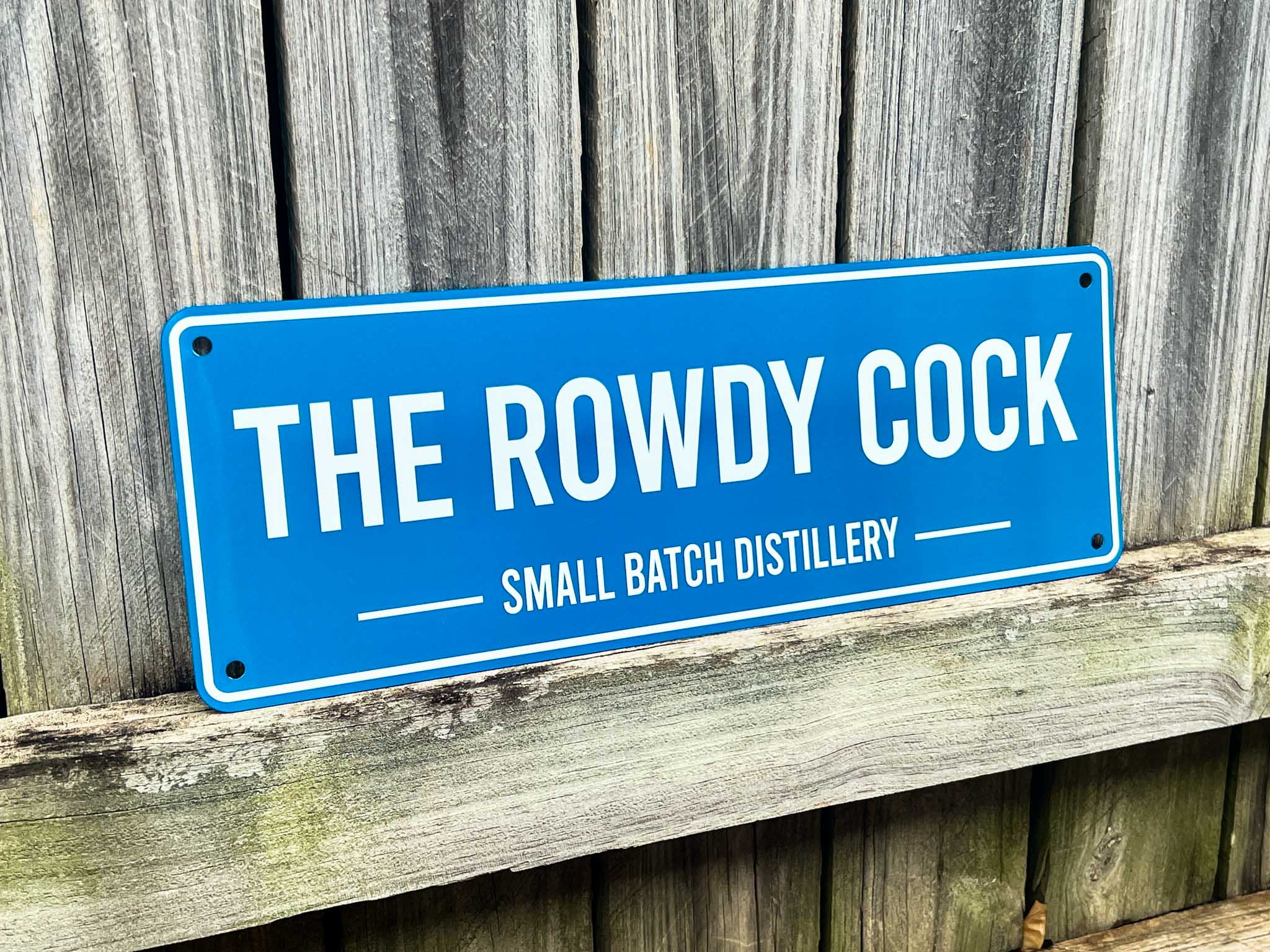 Blue sign with 'The Rowdy Cock Small Batch Distillery' text on a wooden background