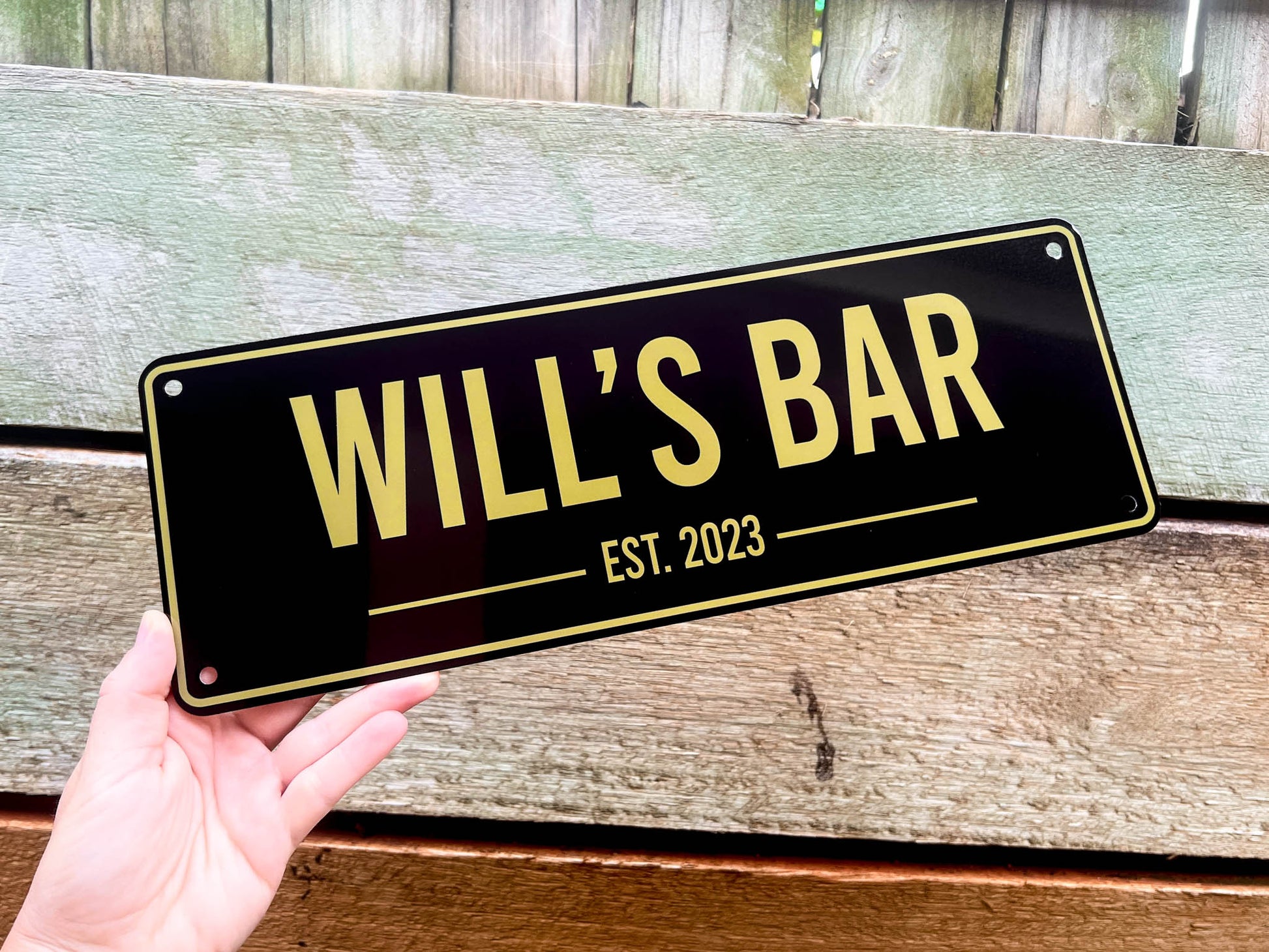 Hand holding a black bar sign with 'WILL'S BAR EST. 2023' in gold letters against a wooden background.