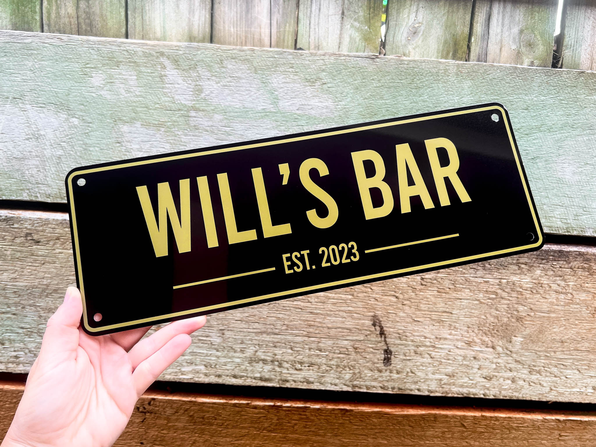 Hand holding a black bar sign with 'WILL'S BAR EST. 2023' in gold letters against a wooden background.