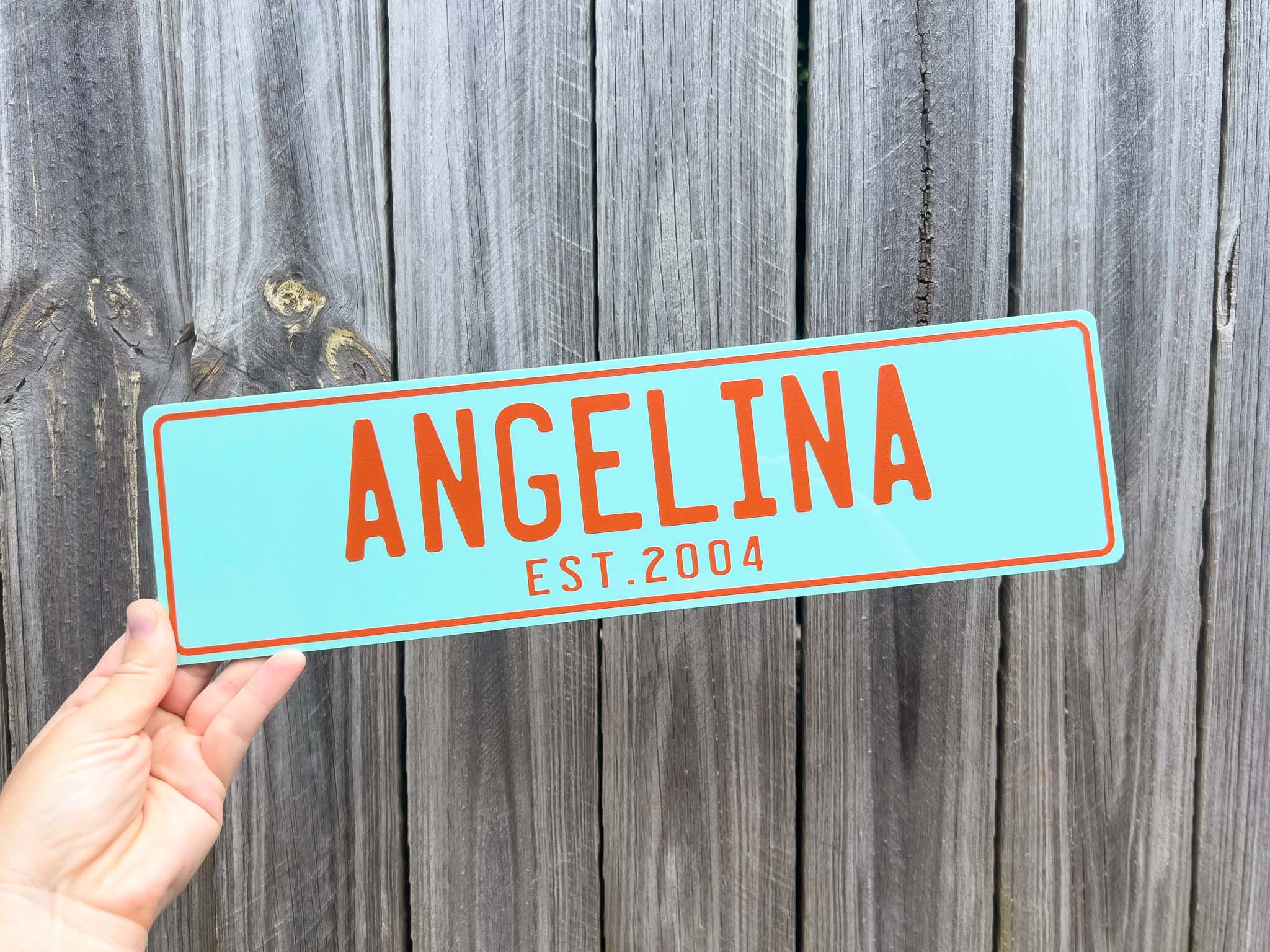Hand holding a turquoise sign with 'ANGELINA EST. 2004' against a wooden background