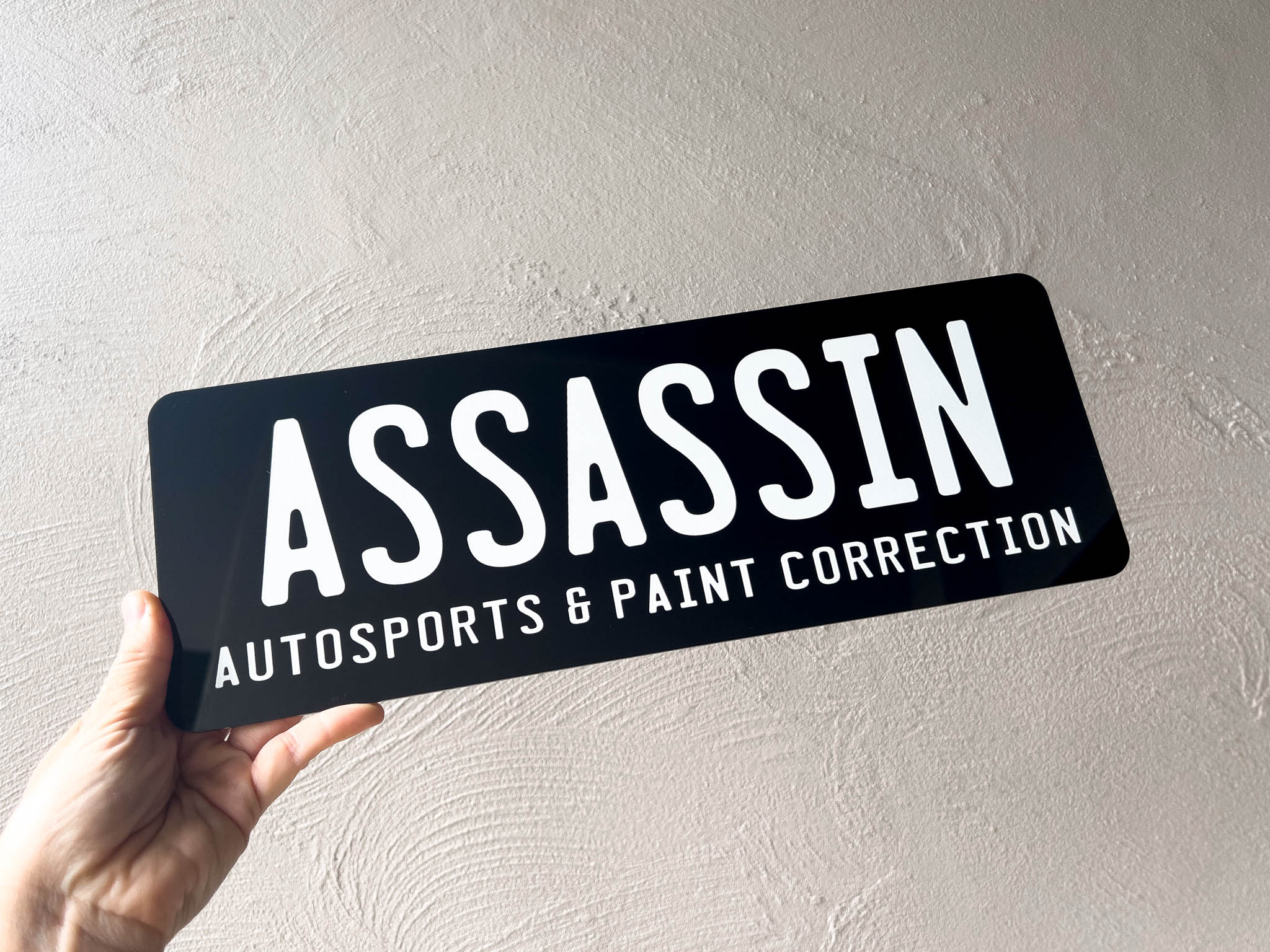 Hand holding a black sign with 'ASSASSIN AUTOSPORTS & PAINT CORRECTION' text against a light grey background