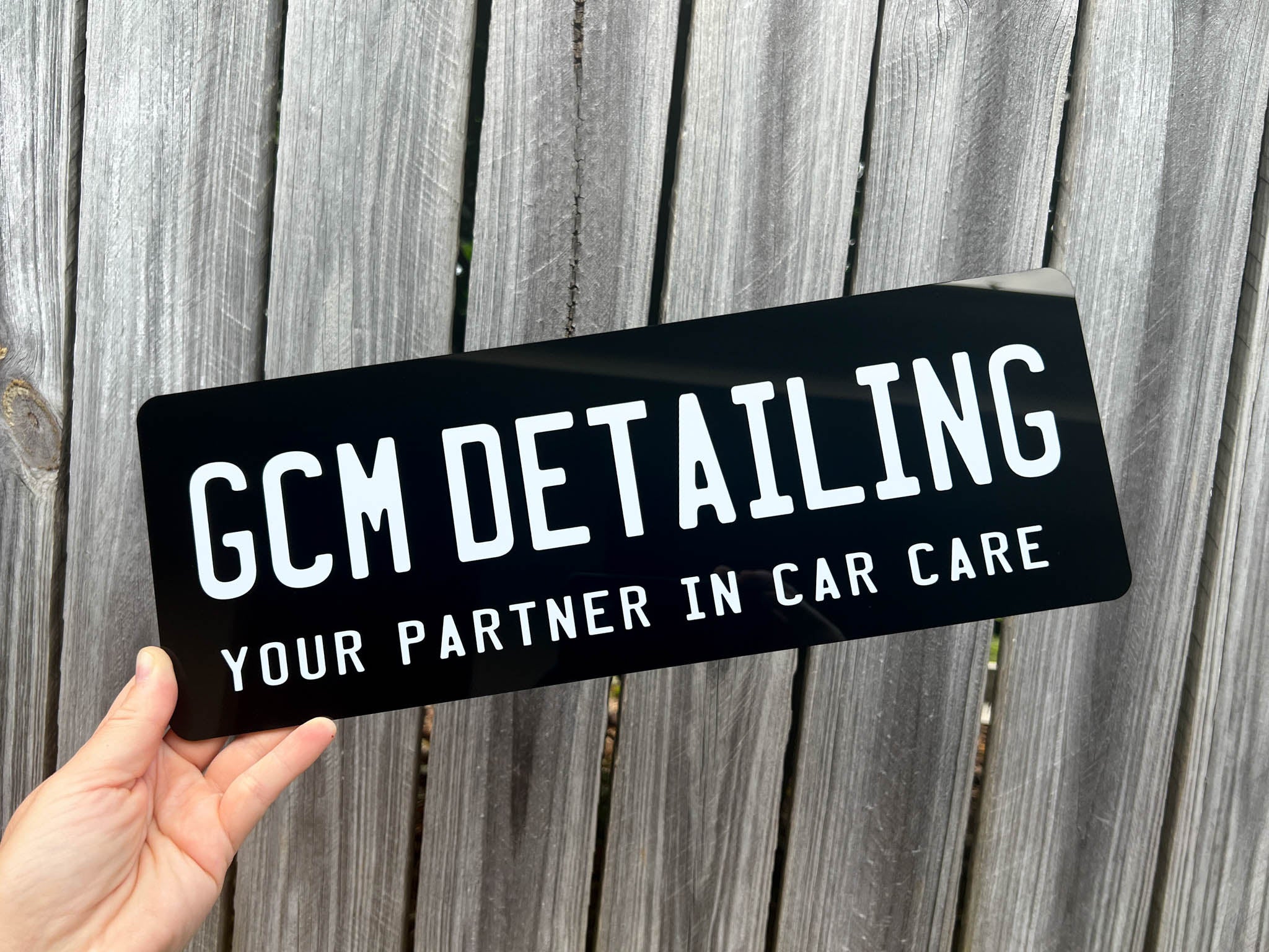 Hand holding a black acrylic plate with 'GCM Detailing' text against a wooden background