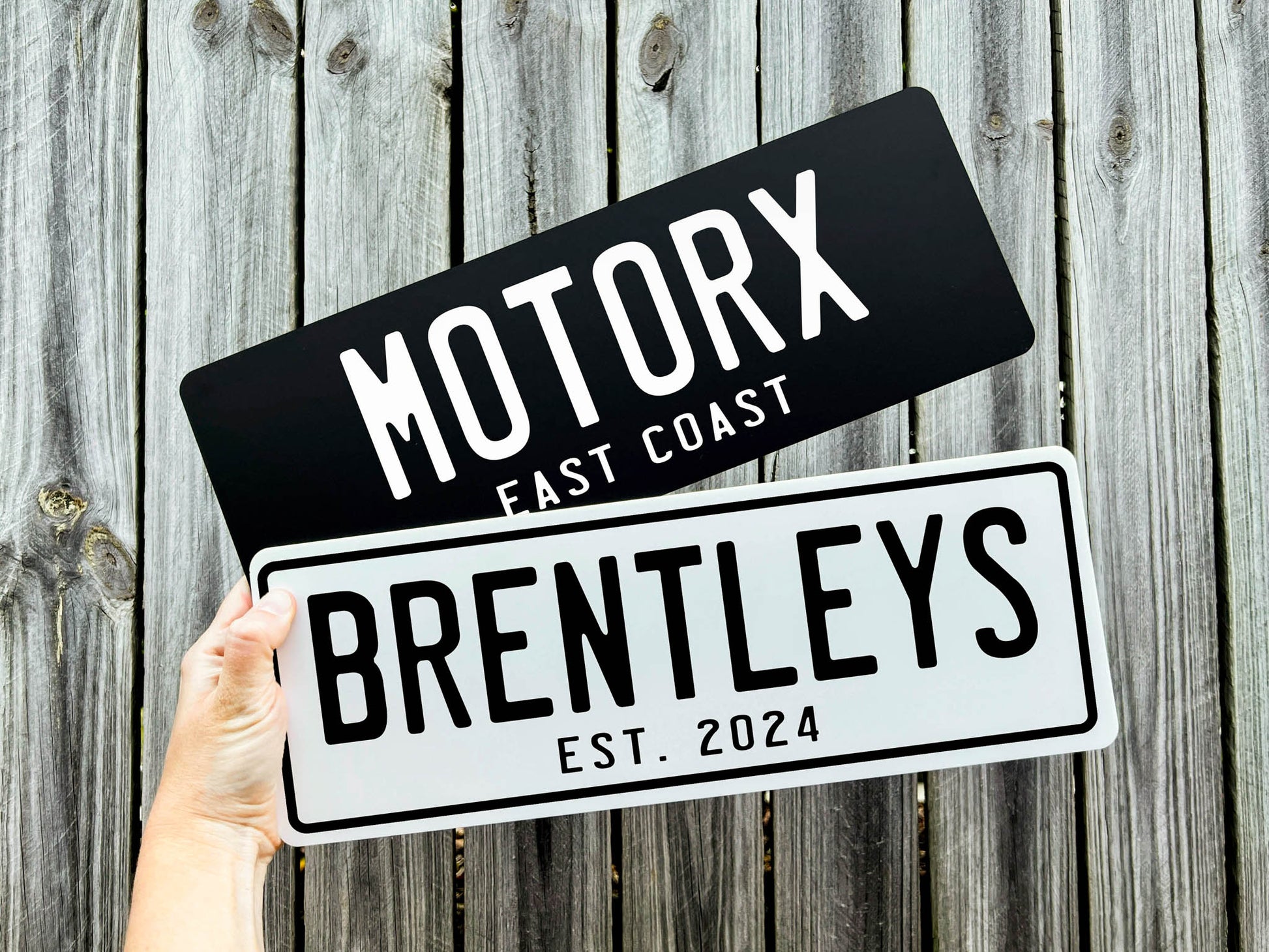 Two custom acrylic cover plates held against a wooden background, one labeled 'Motorx East Coast' and the other 'Brentleys Est. 2024'.