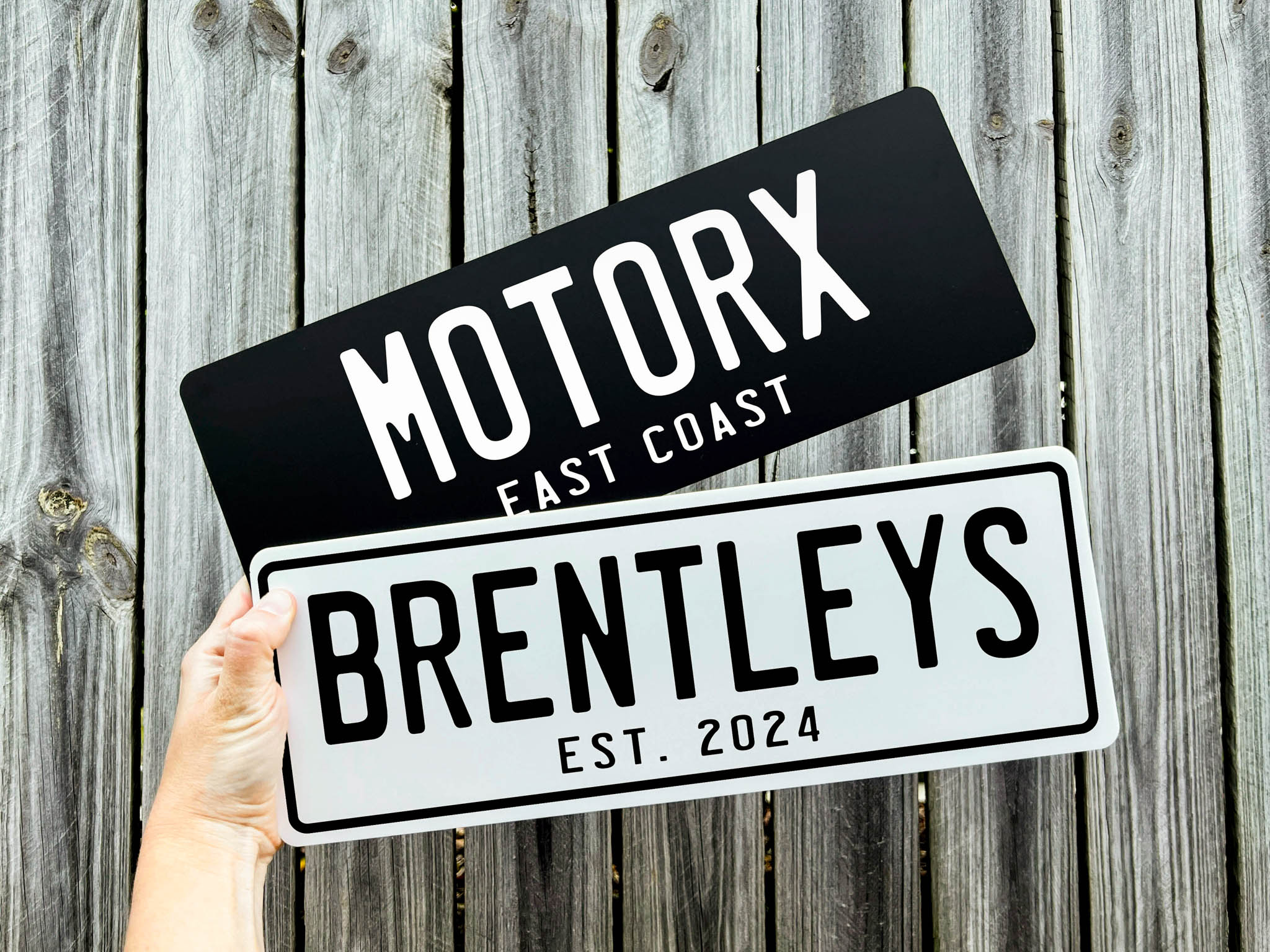 Two custom acrylic cover plates held against a wooden background, one labeled 'Motorx East Coast' and the other 'Brentleys Est. 2024'.