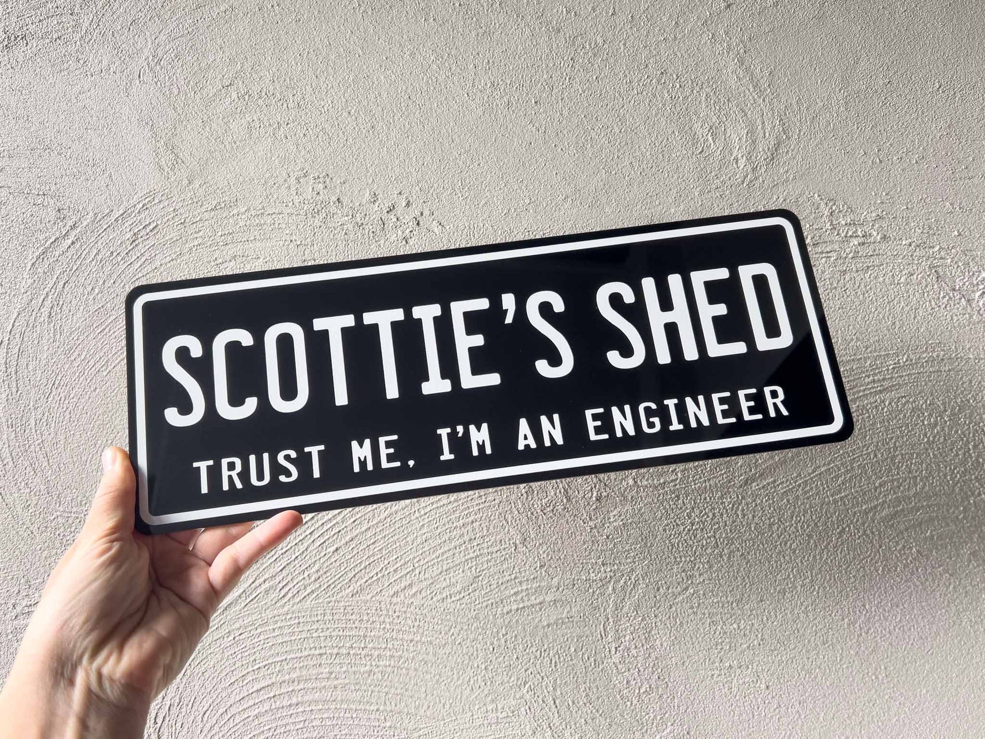 Hand holding a sign that reads 'Scottie's Shed Trust Me, I'm an Engineer' against a textured grey background.