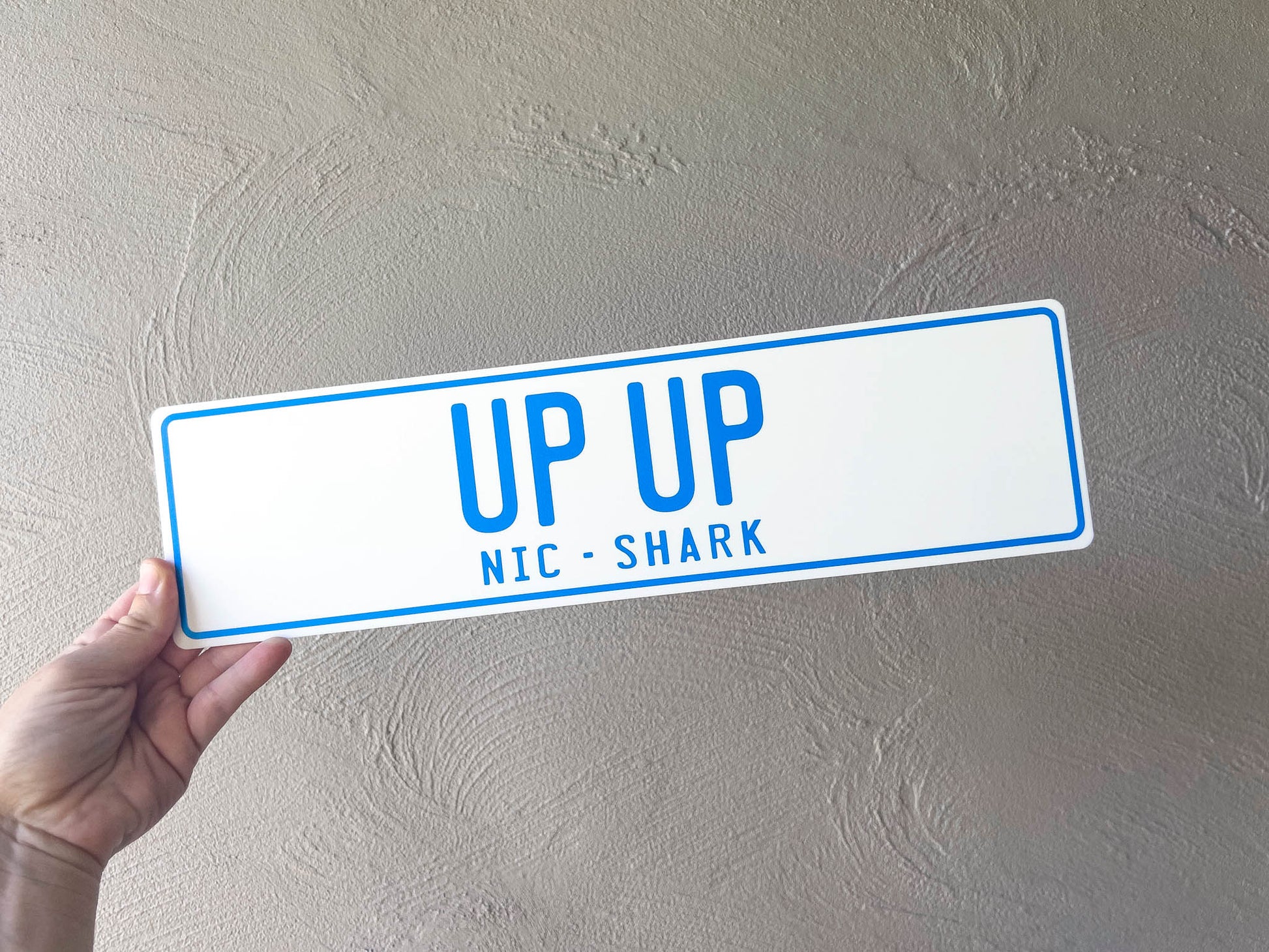 Hand holding a rectangular acrylic slim plate with 'UP UP NIC SHARK' text against a plain background