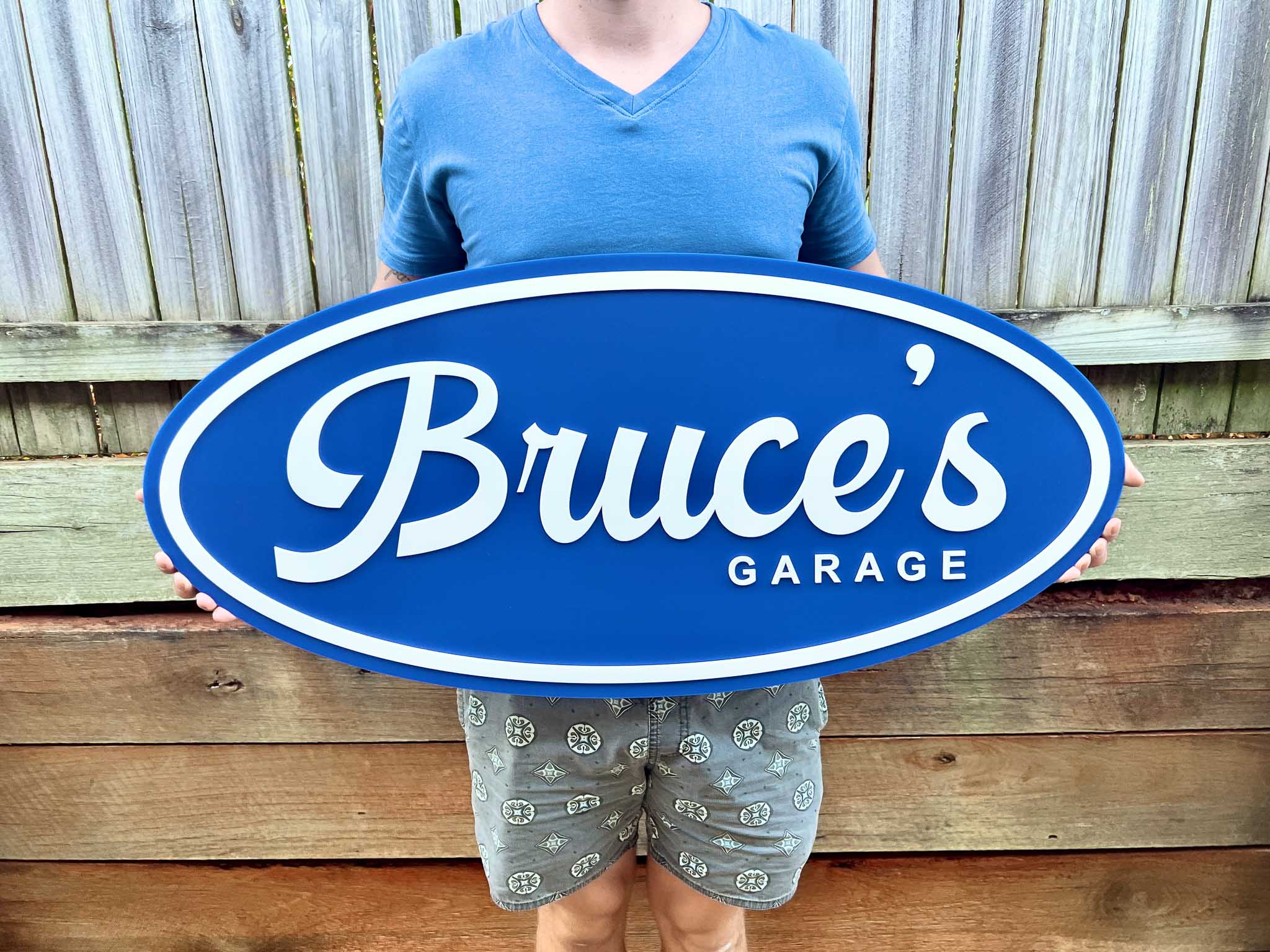 Person holding a blue retro oval sign with 'Bruce's Garage' text against a wooden fence background