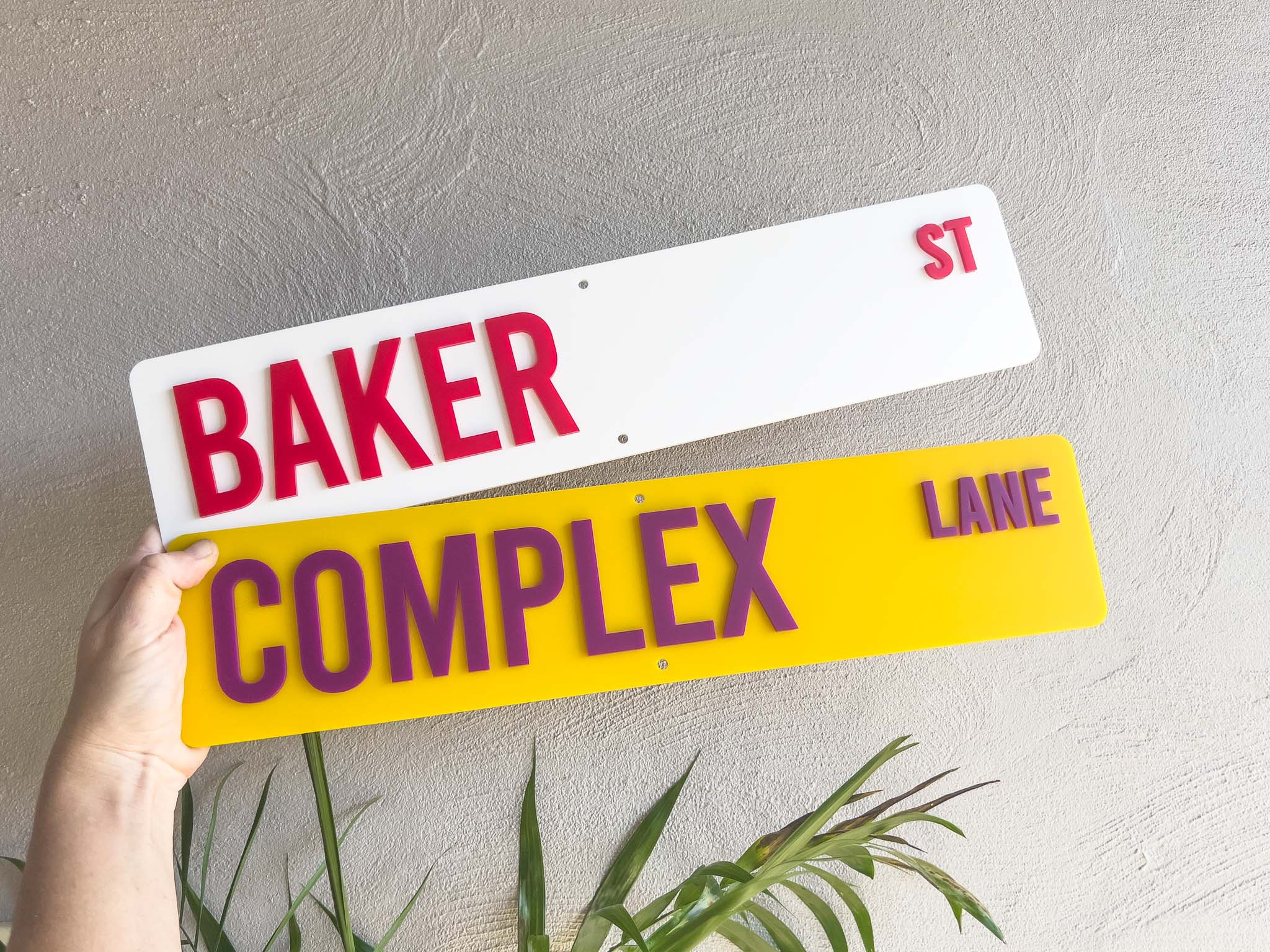 Two street signs, one white with red text 'BAKER ST' and one yellow with purple text 'COMPLEX LANE', held against a light grey wall.