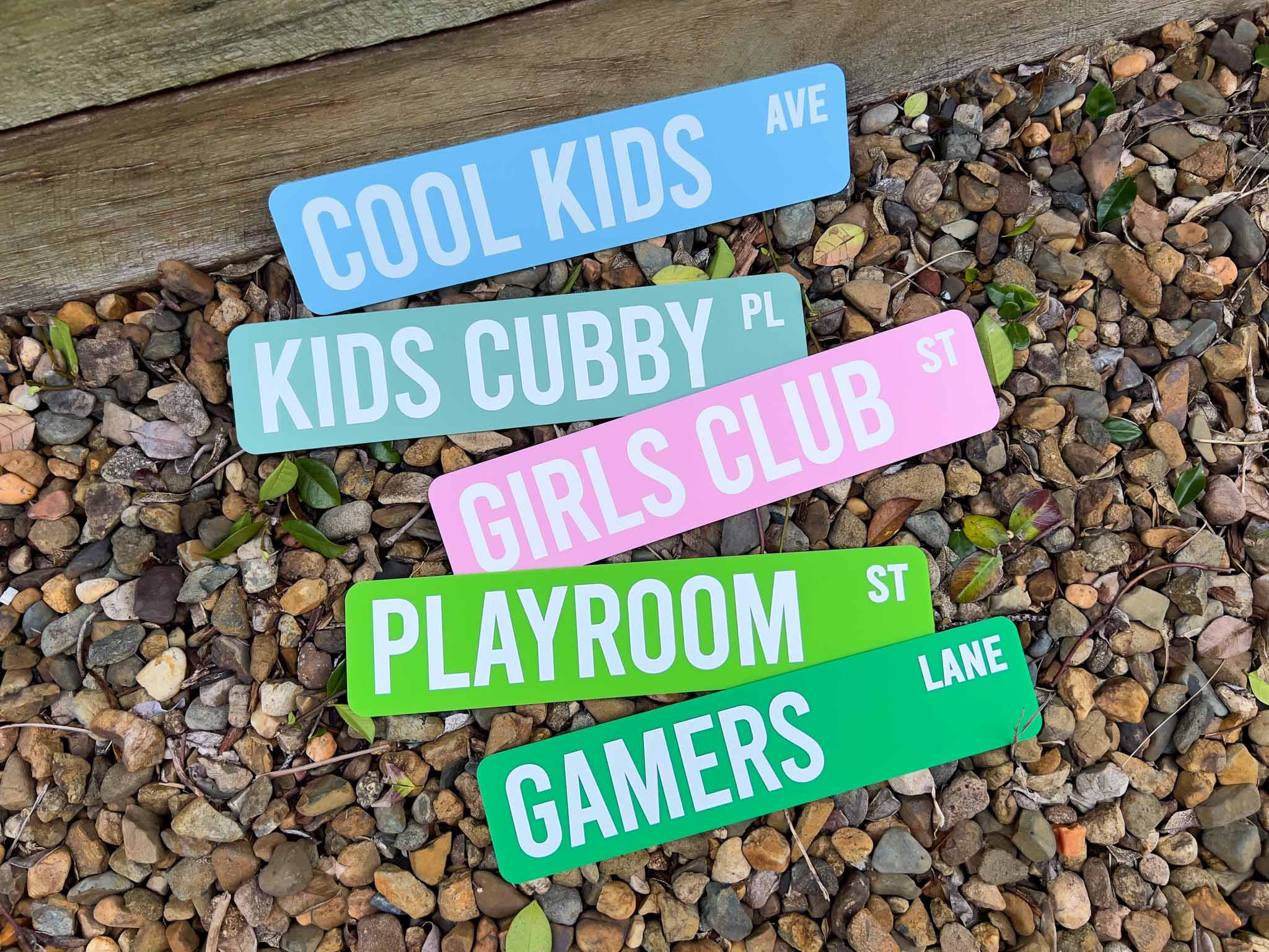 Five colorful street sign plaques with white text are arranged diagonally on a bed of small brown and gray pebbles. The signs read: 'COOL KIDS AVE', 'KIDS CUBBY PL', 'GIRLS CLUB ST', 'PLAYROOM ST', and 'GAMERS LANE'.