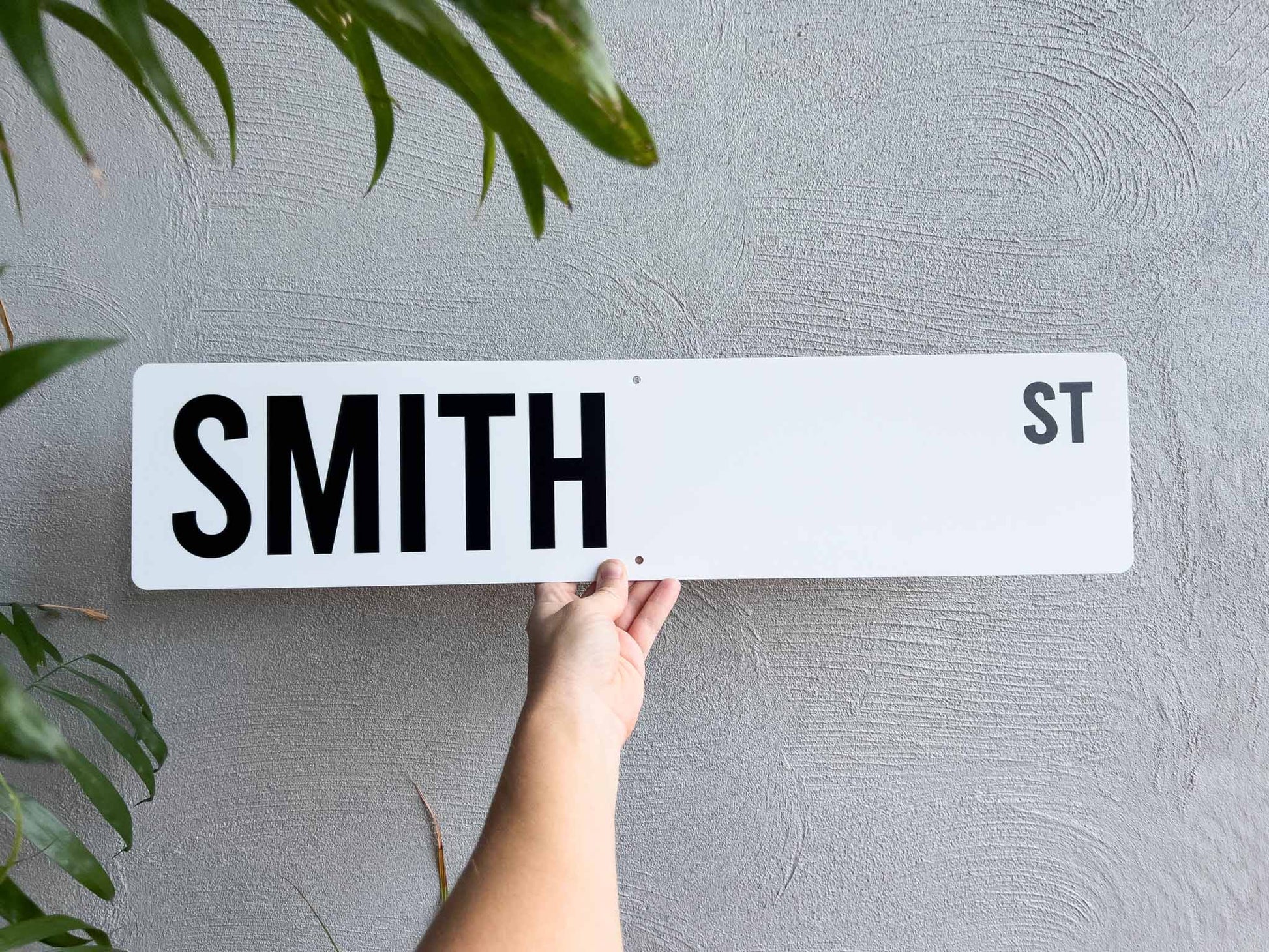 Hand holding a white and black street sign with 'SMITH ST' against a grey wall.