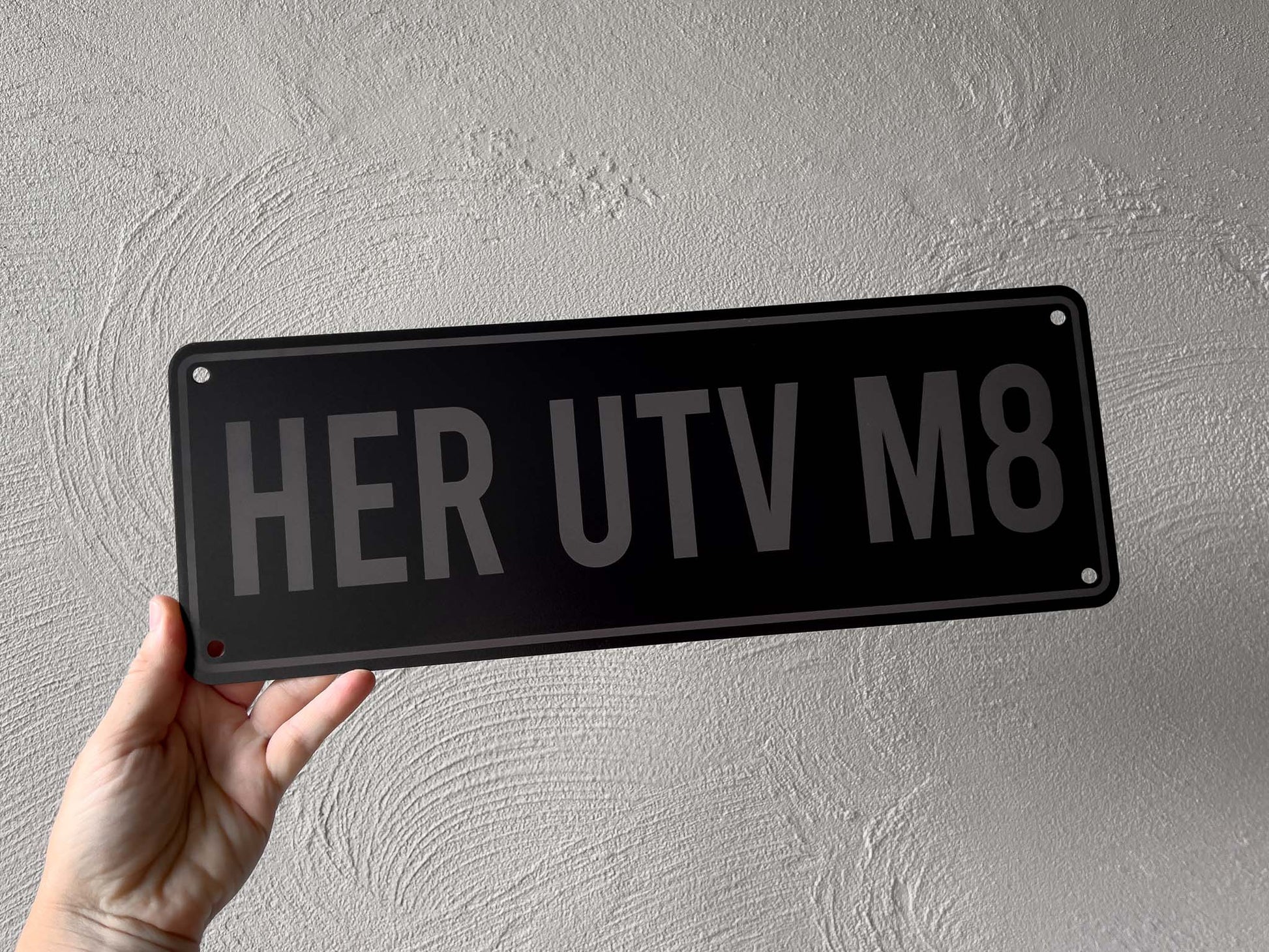 Person holding a black aluminium sign with 'HER UTV M8' text against a grey background