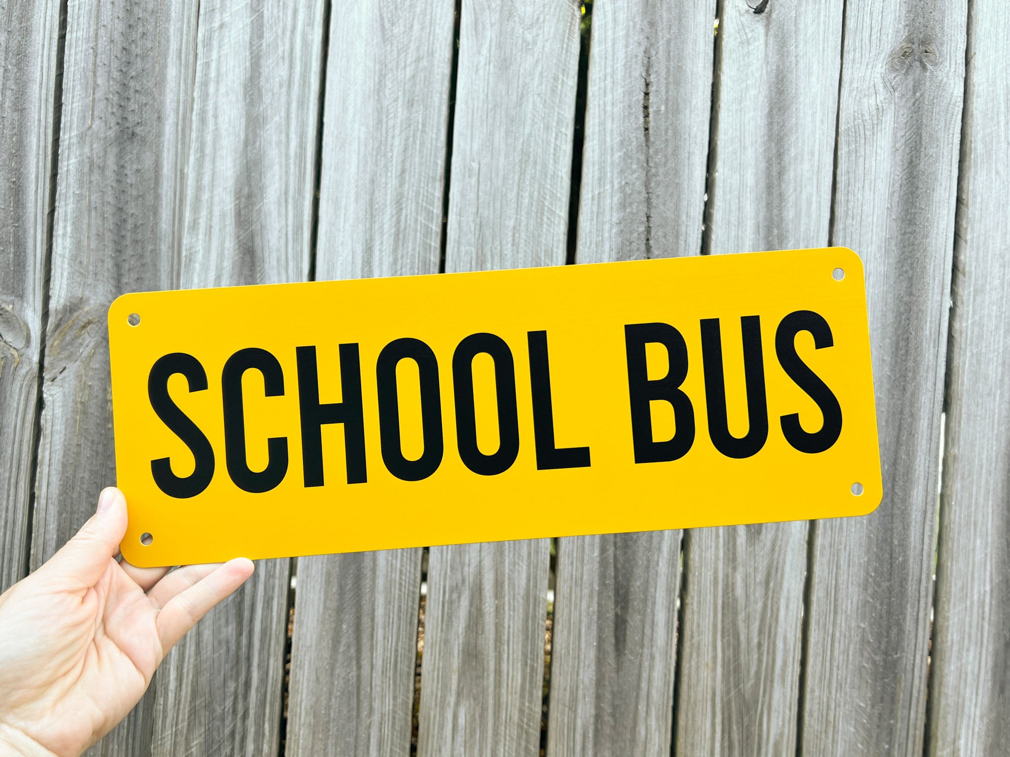 Hand holding a yellow 'SCHOOL BUS' sign against a wooden fence background