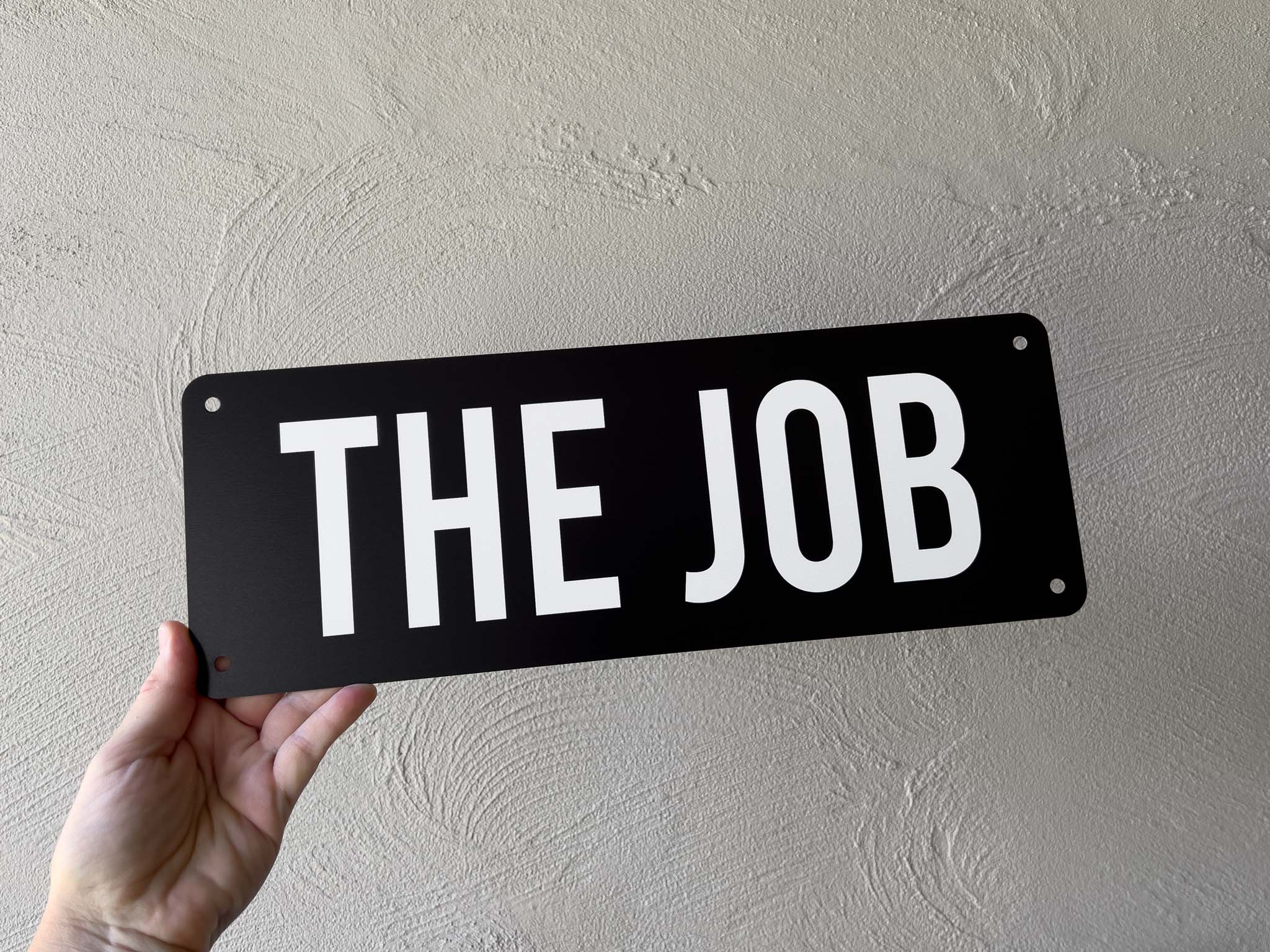 Hand holding a black metal sign with 'THE JOB' in white letters against a light grey wall.