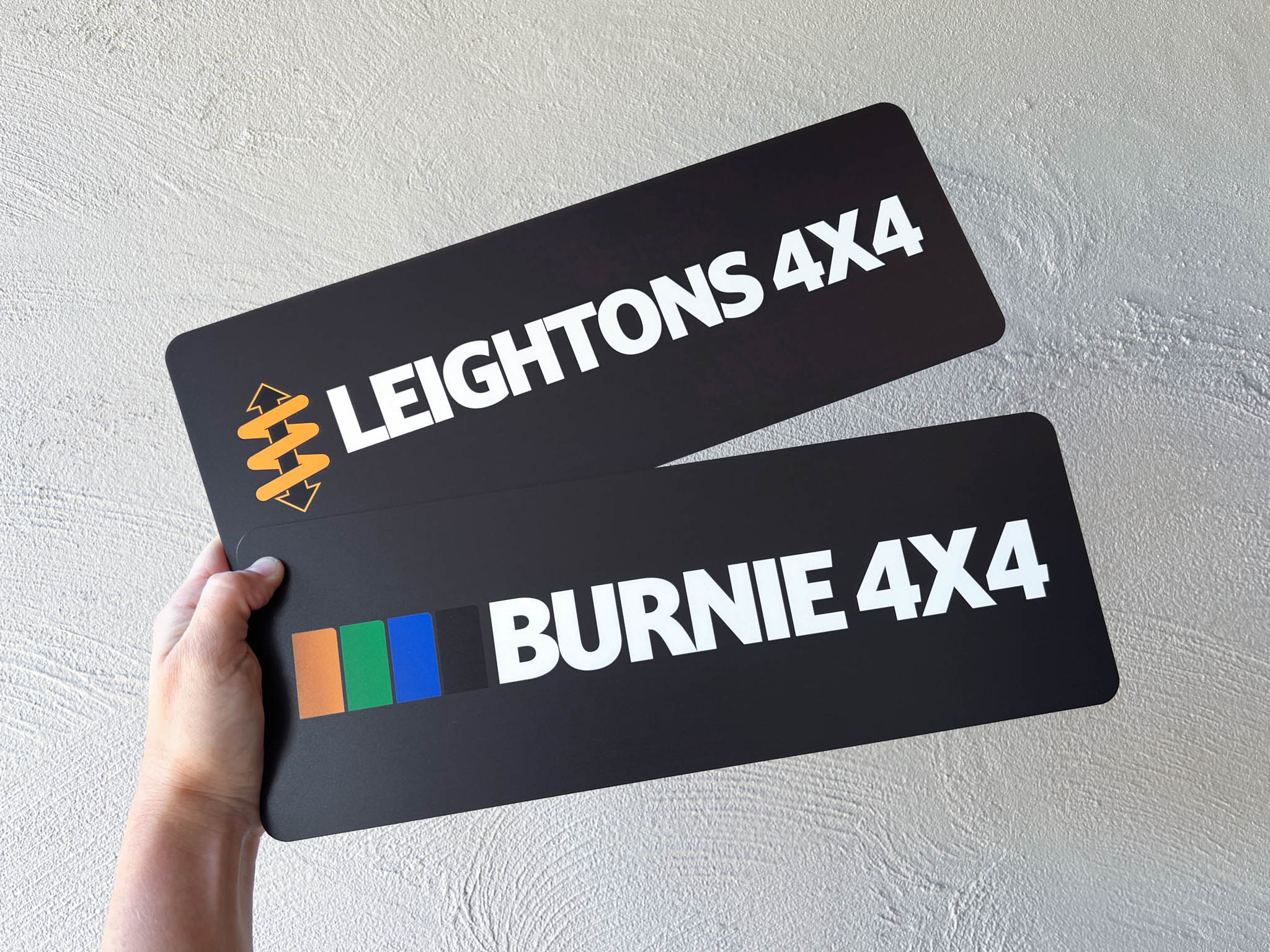 Two black license plates with 'Leightons 4X4' and 'Burnie 4X4' branding held by a hand against a light grey background.