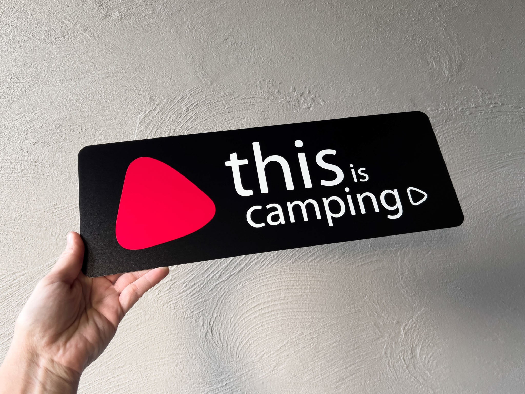Hand holding a black sign with 'this is camping' and a red play button symbol on a light grey background.