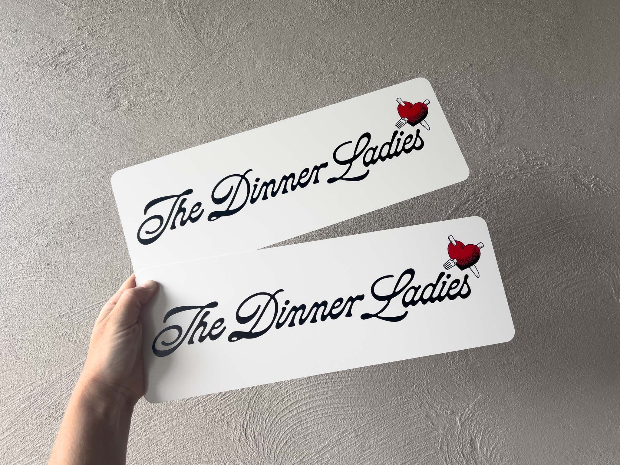 Two 'The Dinner Ladies' signs with a hand holding one of them against a light grey background.