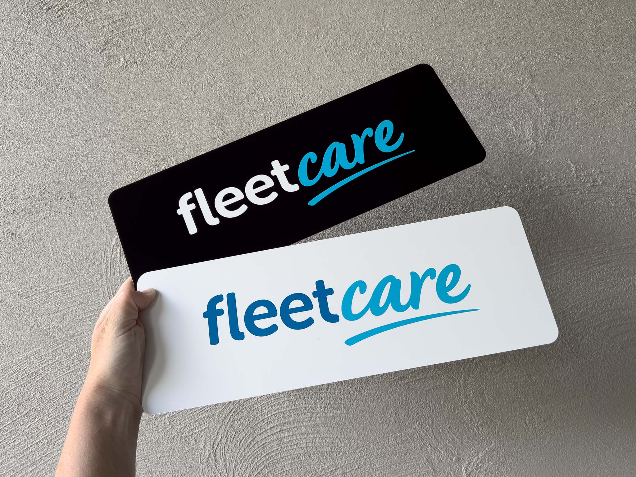 Two 'fleetcare' custom printed logo signs, one black and one white, held against a grey background.