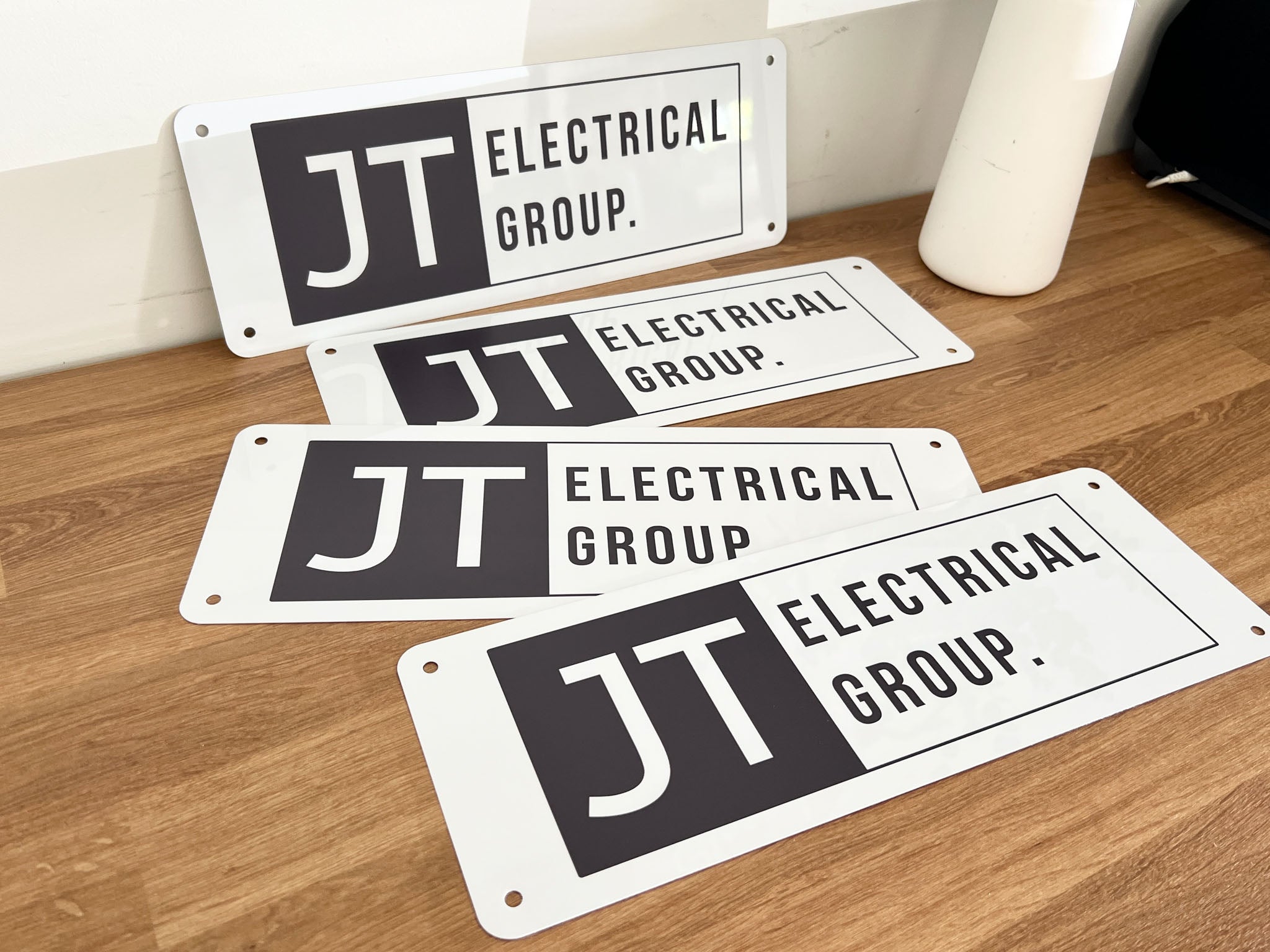 Four metal signs with 'JT Electrical Group' logo on a wooden surface.