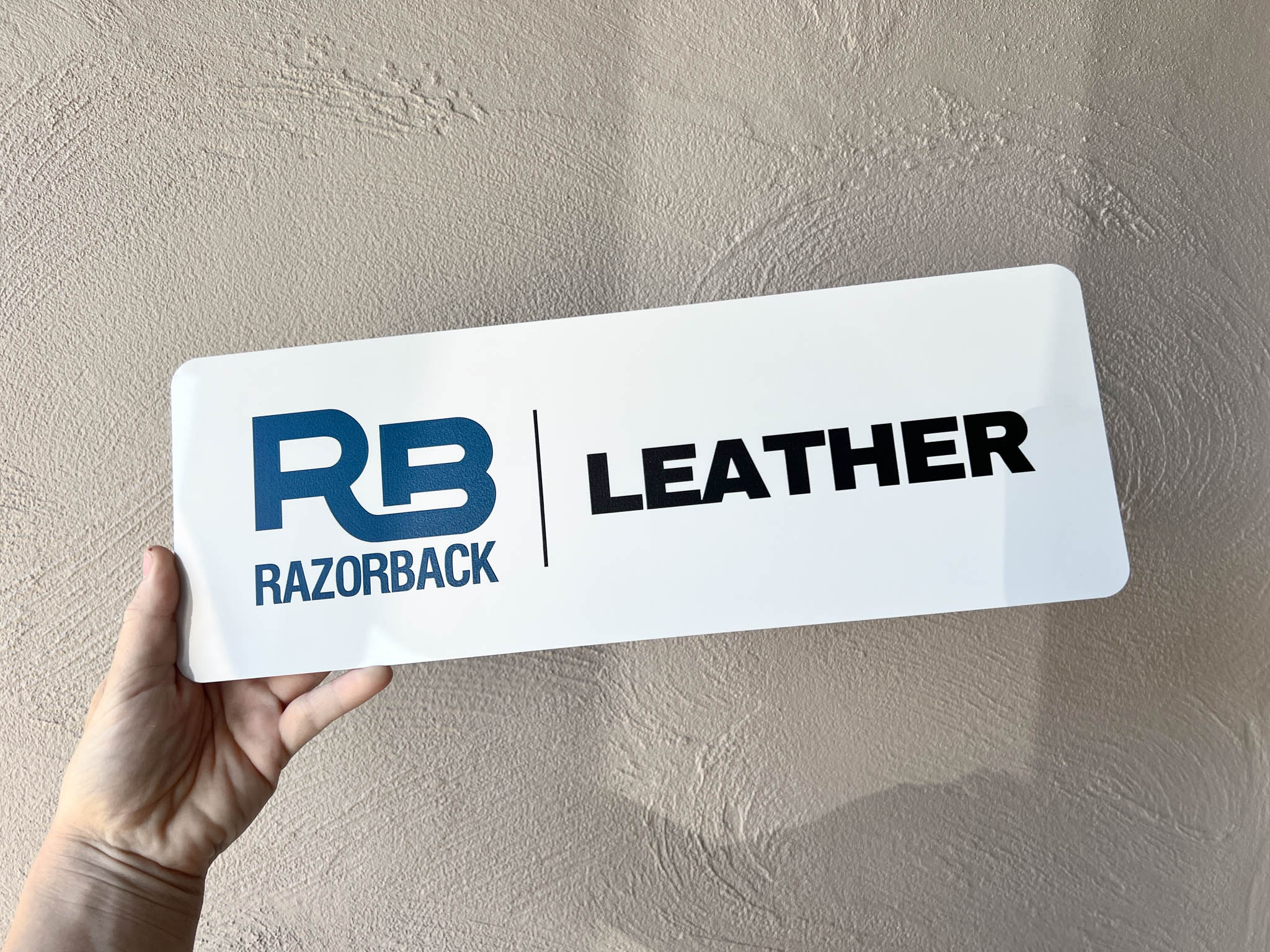 Hand holding a custom printed logo sign with 'Razorback Leather' branding against a beige wall.