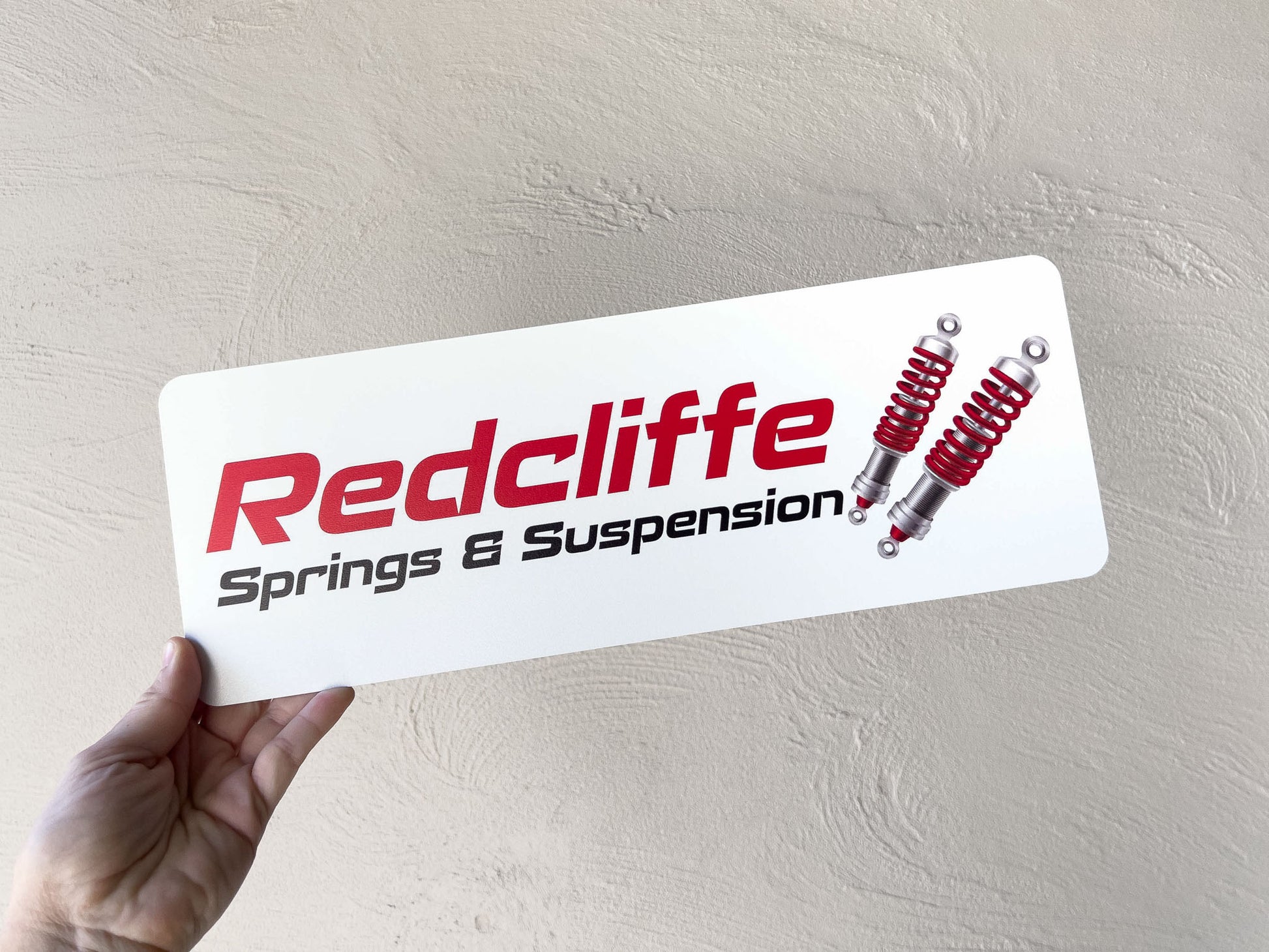 Hand holding a Redcliffe Springs & Suspension metal plate with suspension parts on a light grey background