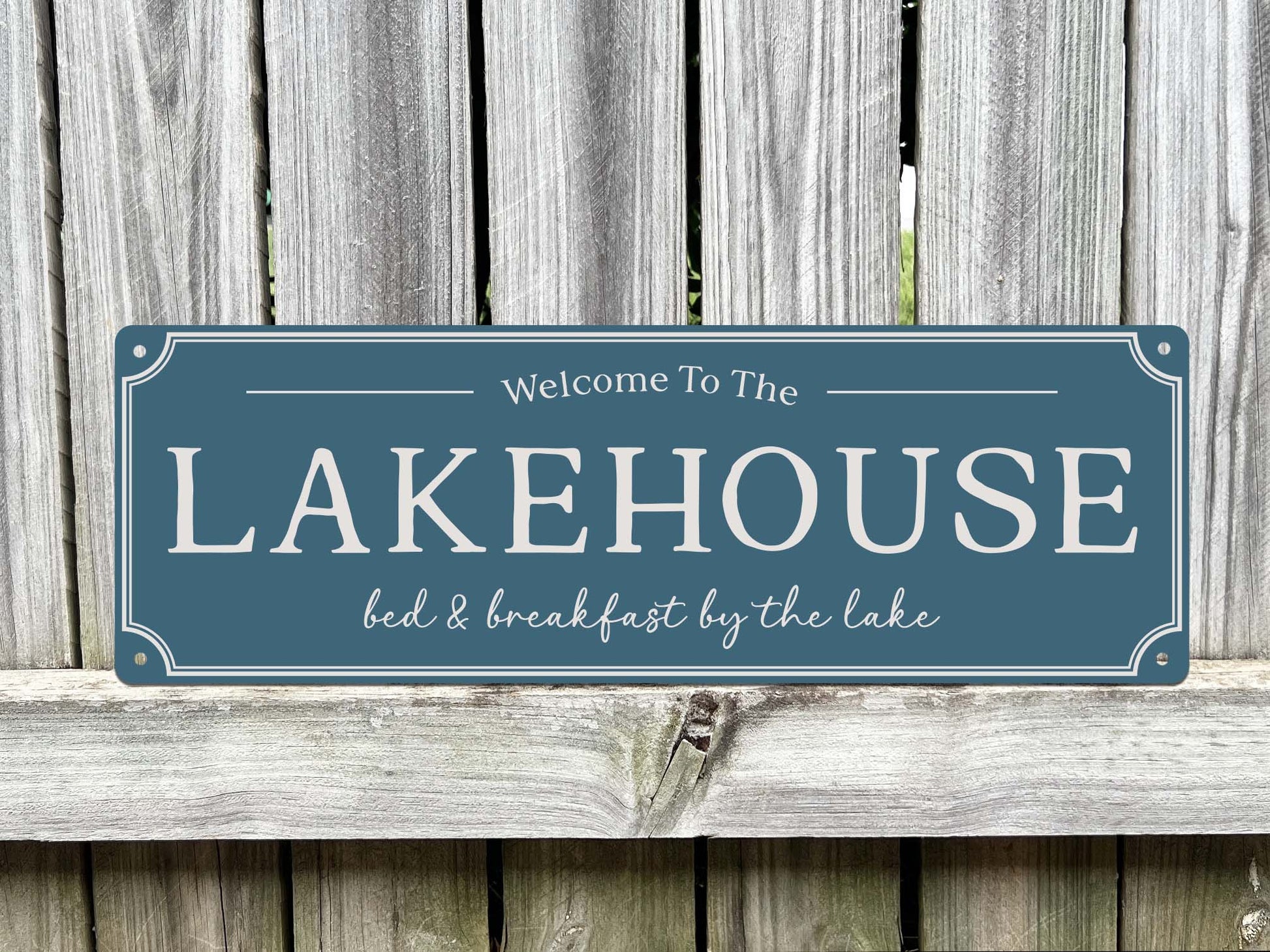 A rectangular farmhouse style sign in a slate blue sign colour with white lettering that reads 'Welcome To The LAKEHOUSE bed & breakfast by the lake,' displayed against a weathered grey wooden fence.