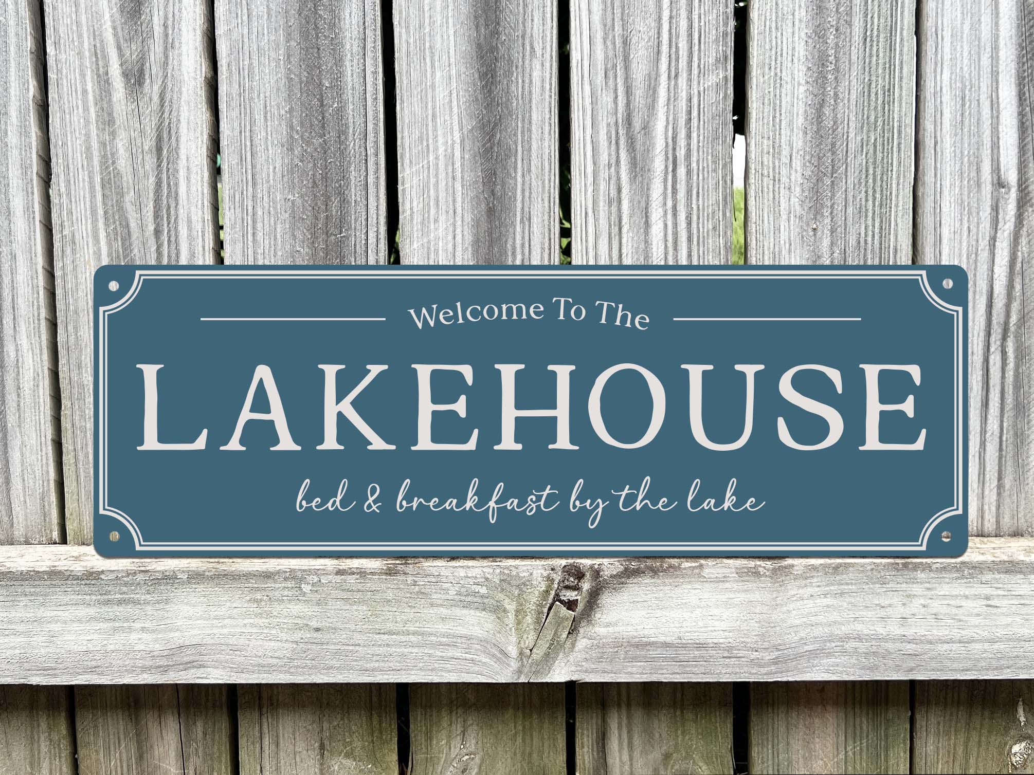 A rectangular farmhouse style sign in a slate blue sign colour with white lettering that reads 'Welcome To The LAKEHOUSE bed & breakfast by the lake,' displayed against a weathered grey wooden fence.