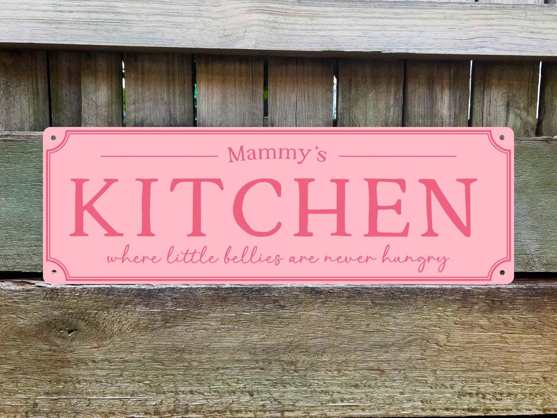 A pink vintage cabin style decorative sign with rounded corners and printed text reading 'Mammy's KITCHEN where little bellies are never hungry,' shown against a weathered wooden fence background.