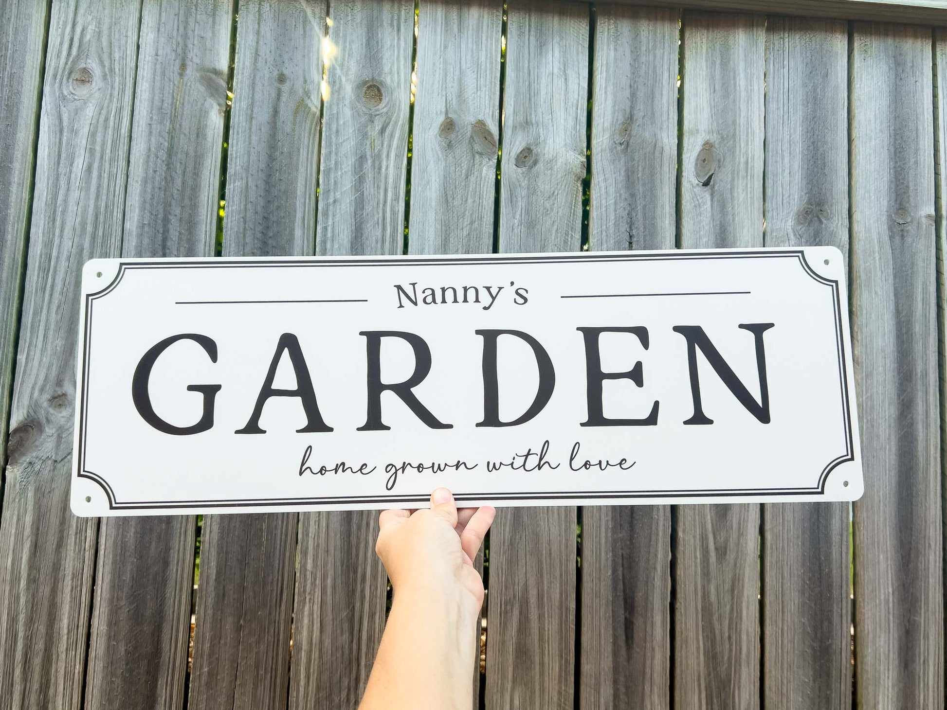 A white rectangular country style sign with black lettering that reads 'Nanny's GARDEN home grown with love,' held by a hand in front of a weathered gray wooden fence.