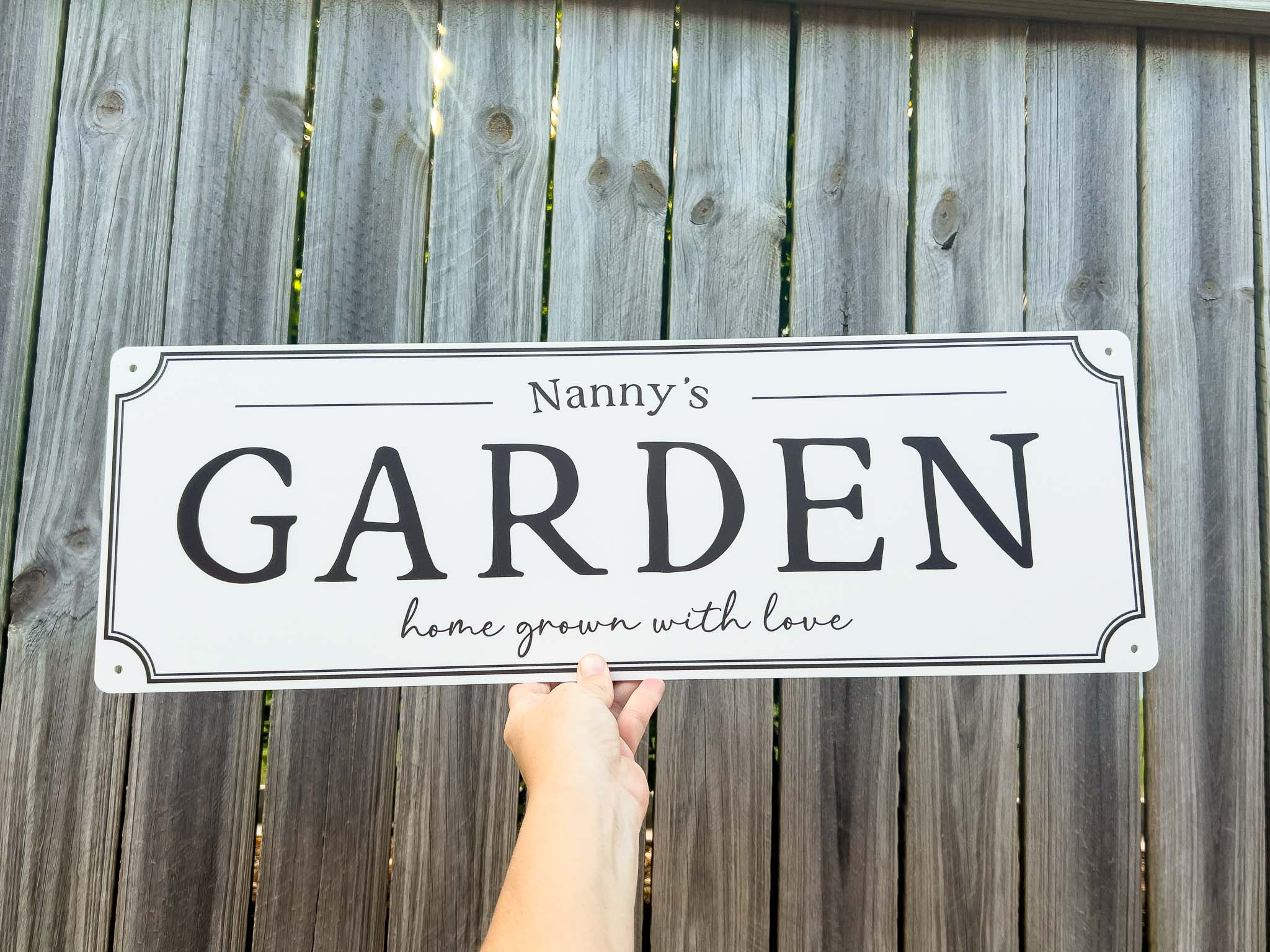 A white rectangular country style sign with black lettering that reads 'Nanny's GARDEN home grown with love,' held by a hand in front of a weathered gray wooden fence.