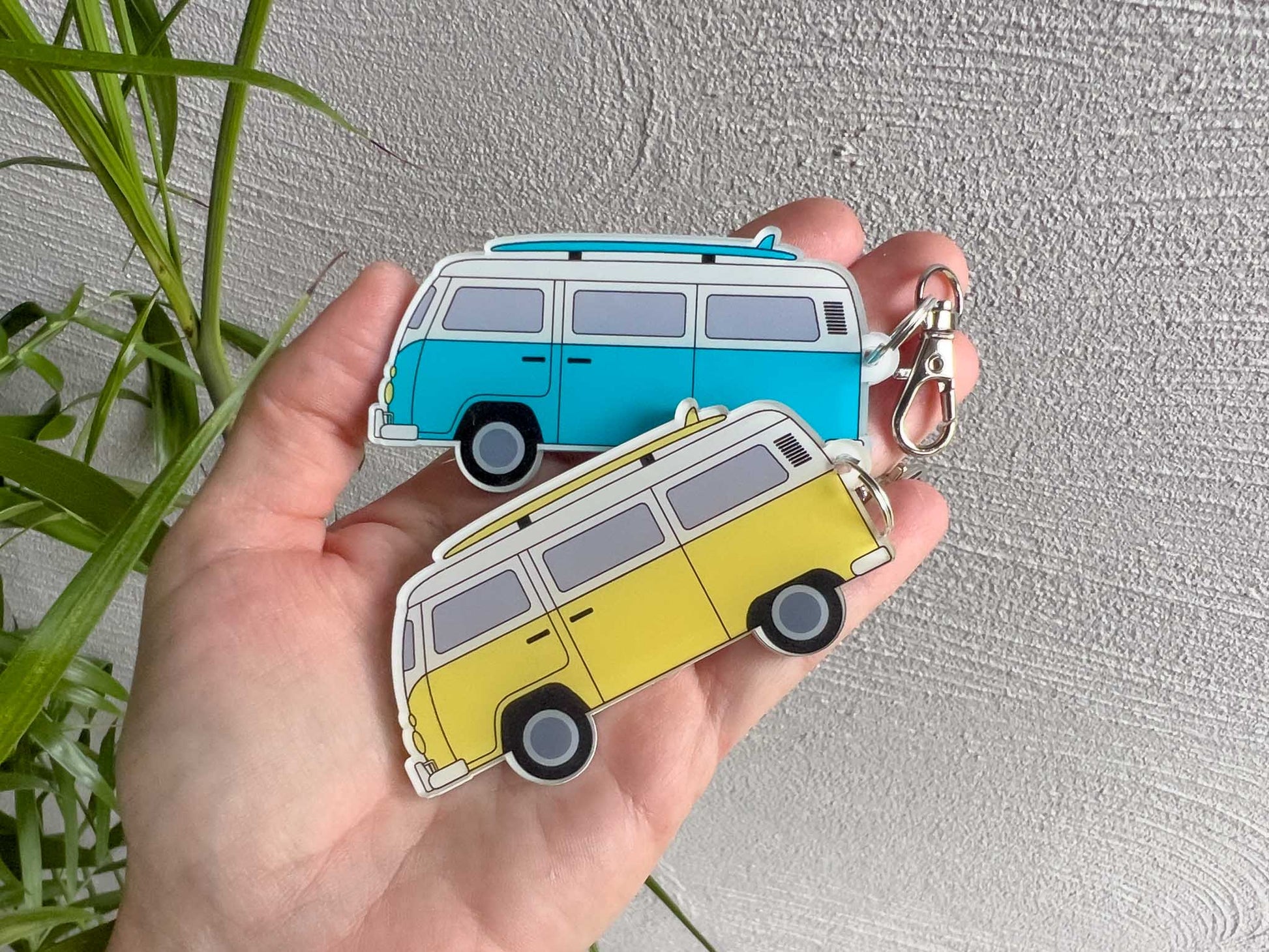 A hand holds two small laser cut, UV printed acrylic keychains shaped like retro campervans, one in light blue and white and one in yellow and white, against a textured light grey wall with green plant leaves at the side.