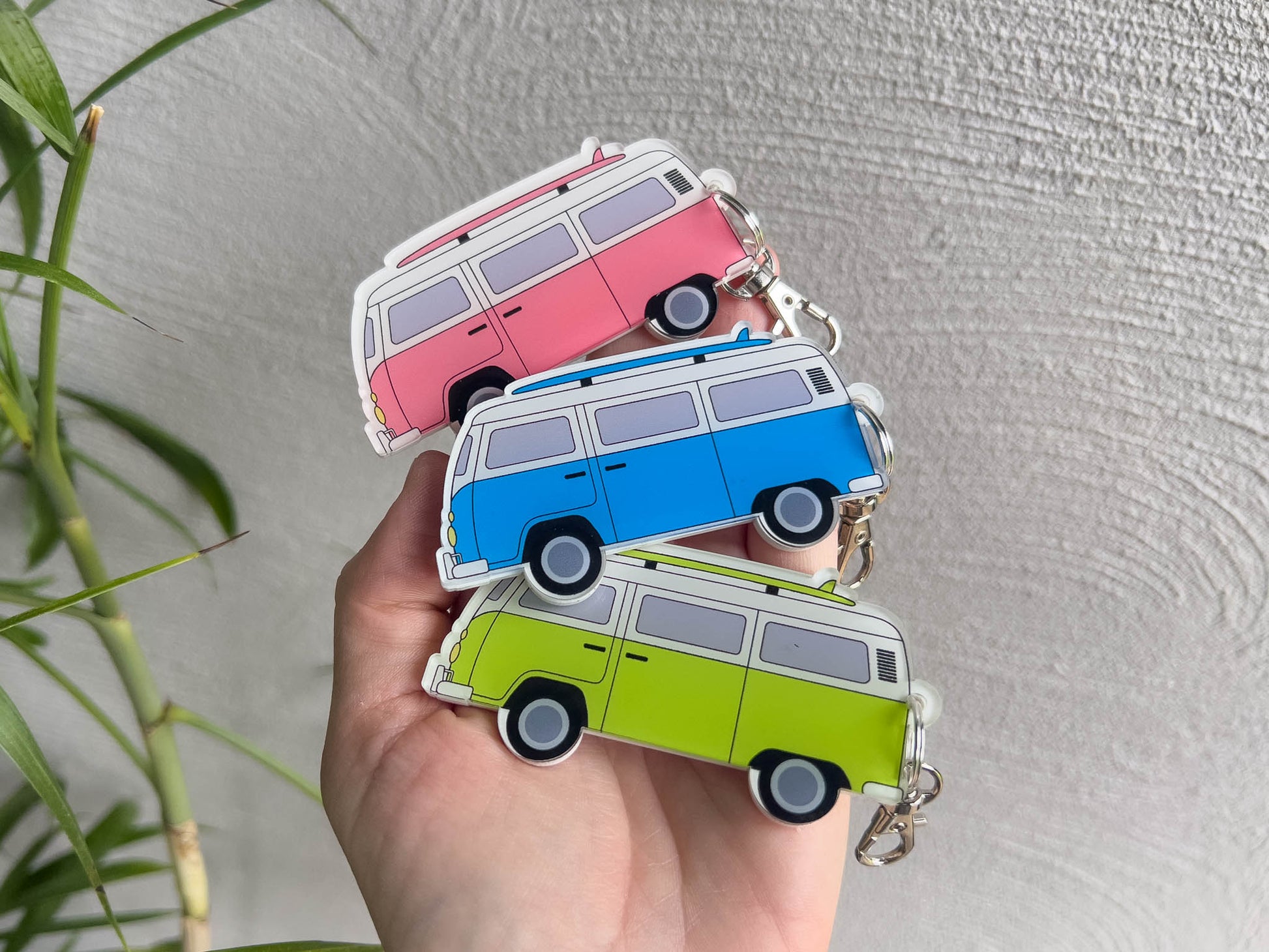 Three small retro-style surf van keychains in pink, blue, and lime green are held in a hand against a textured light grey wall, next to a palm tree.