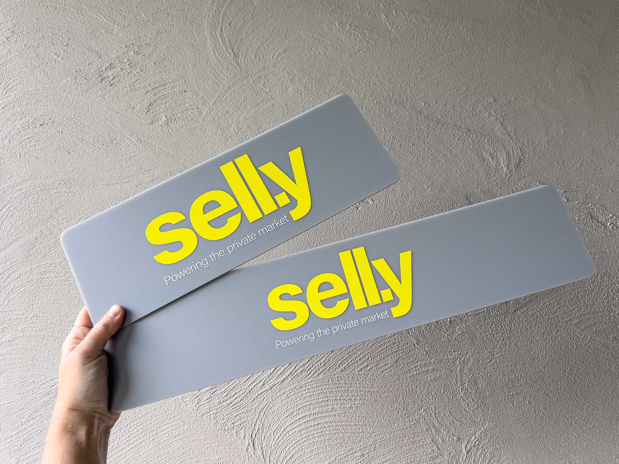 Two custom printed grey acrylic logo plates with the brand name 'Sell.y' printed in yellow and white.
