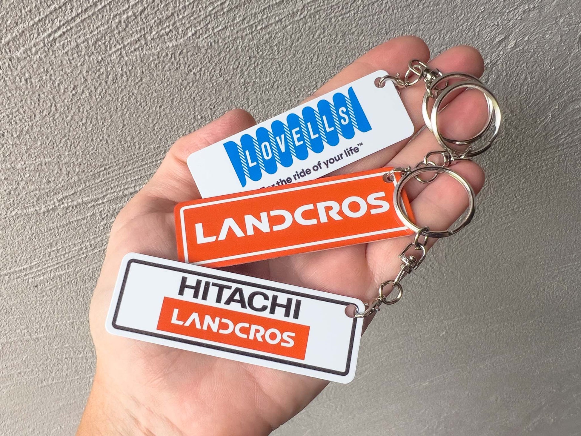 A hand holding three rectangular aluminium keychains with metal key rings. The keychains feature Landcros branding in white, orange, and black, with one keychain also showing the blue Lovells logo.