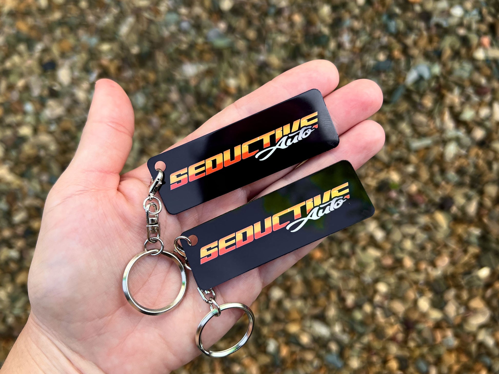 Two black rectangular aluminium keychains with 'SEDUCTIVE Auto' branding in orange, yellow, and white, held in a hand against a gravel background.