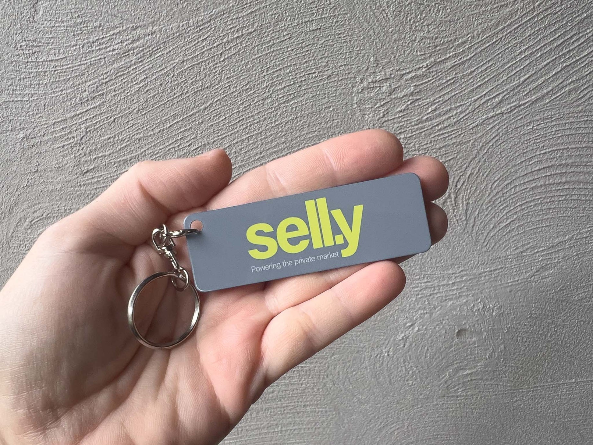 A hand holding a grey rectangular aluminium keychain with the brand name 'Sell.y' in bright yellow and a silver split keyring, set against a textured gray wall.