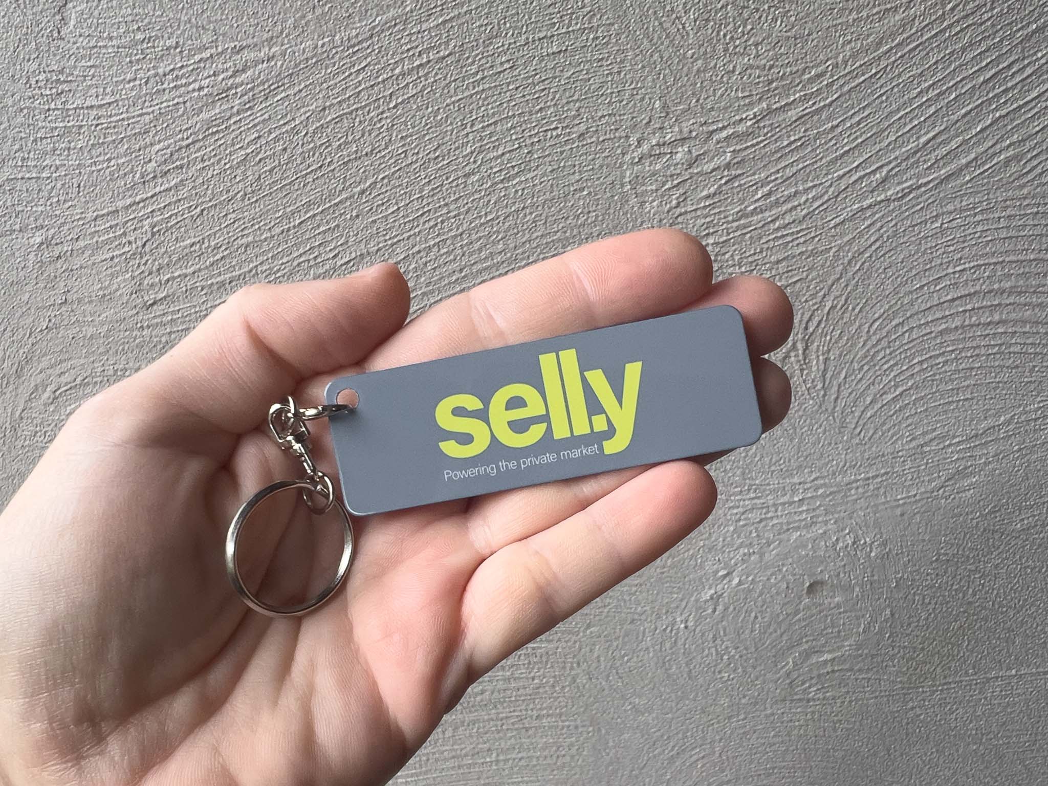 A hand holding a grey rectangular aluminium keychain with the brand name 'Sell.y' in bright yellow and a silver split keyring, set against a textured gray wall.