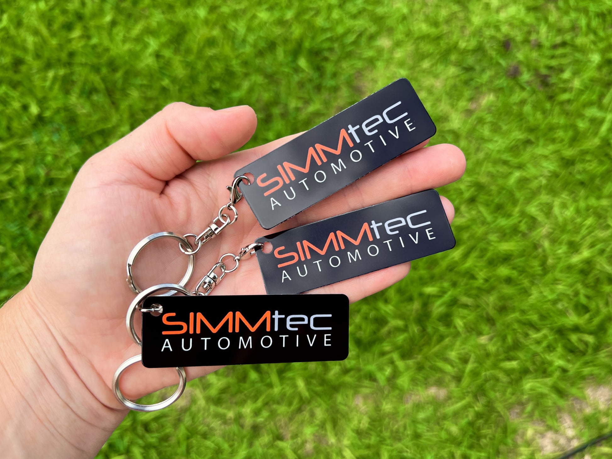 Three black Aluminium SIMMtec Automotive logo keychains held in a hand against a green grass background.